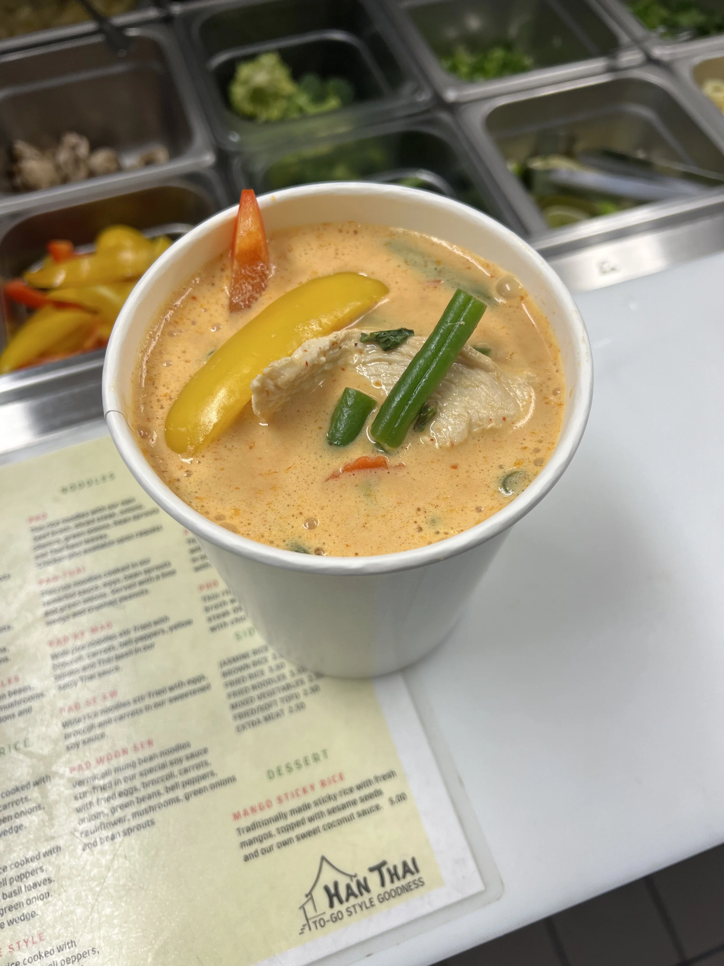 A cup of Thai curry with chicken, green beans, yellow pepper, and red pepper slices, placed on a menu at a restaurant. In the background, there are containers with various chopped vegetables and ingredients.