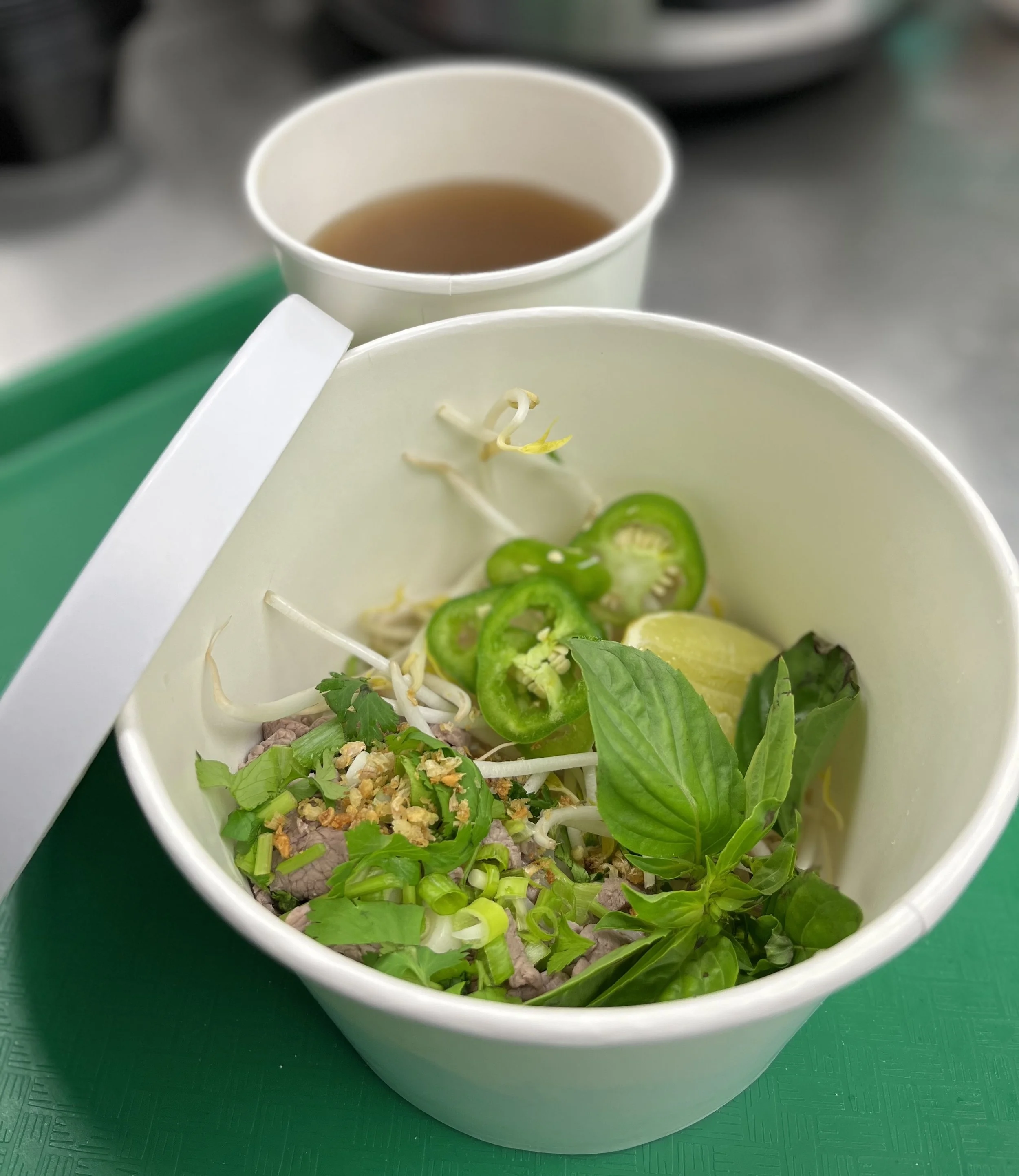 Inside a white takeout bowl, there are fresh green vegetables, bean sprouts, sliced jalapeño peppers, lemon wedges, and chopped nuts. In the background, there is a cup of hot tea or light broth on a green surface.