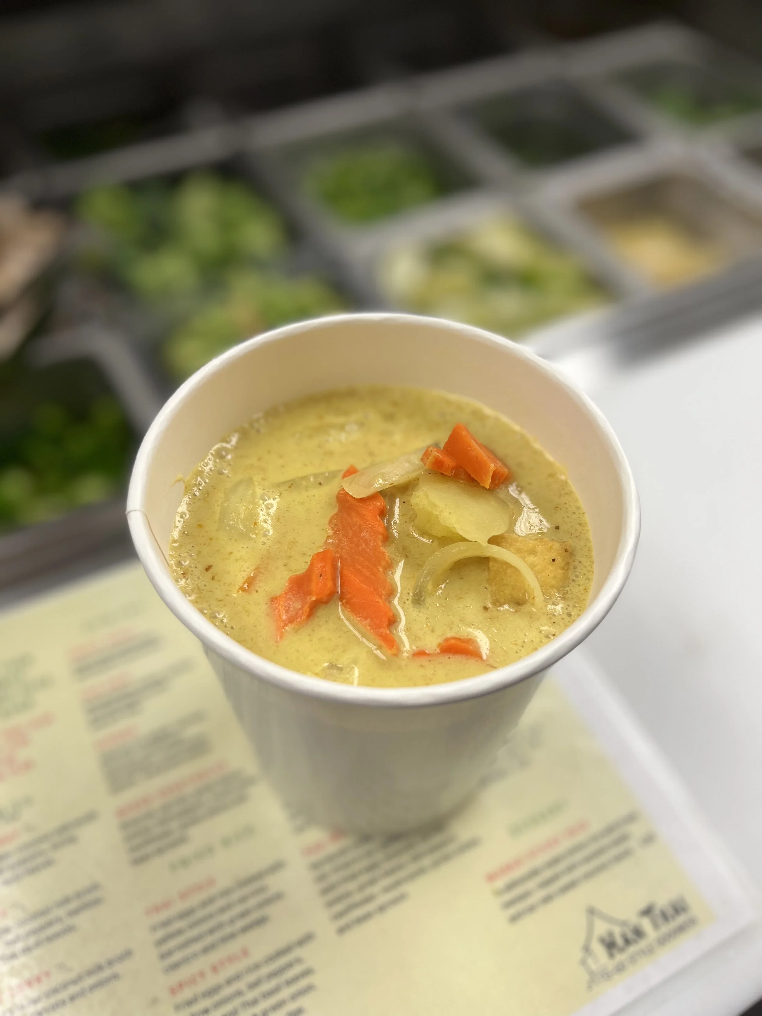 A cup of creamy chicken noodle soup with visible carrots and onions, placed on a paper menu or food brochure.