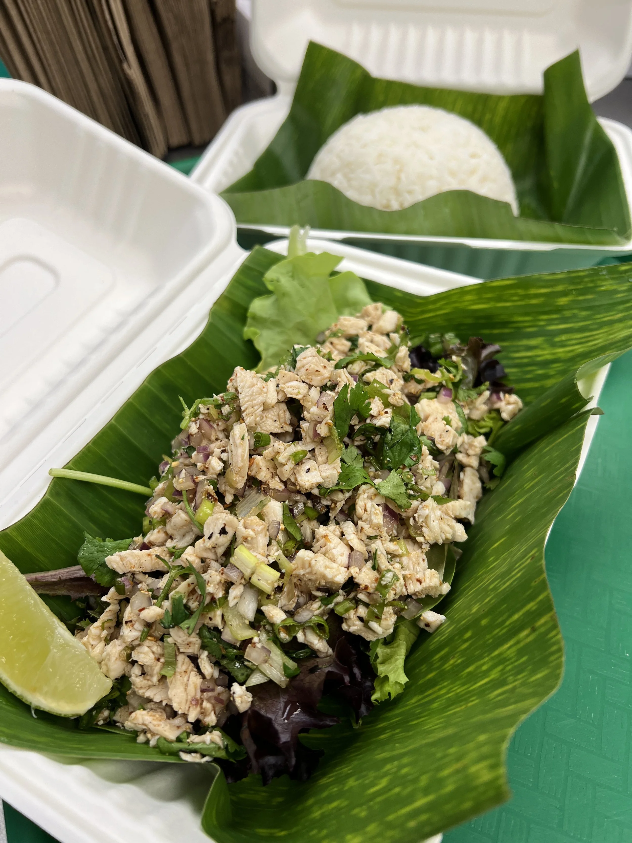 Close-up of a chicken salad on a green banana leaf, with lime wedges on the side, in a white takeout container.