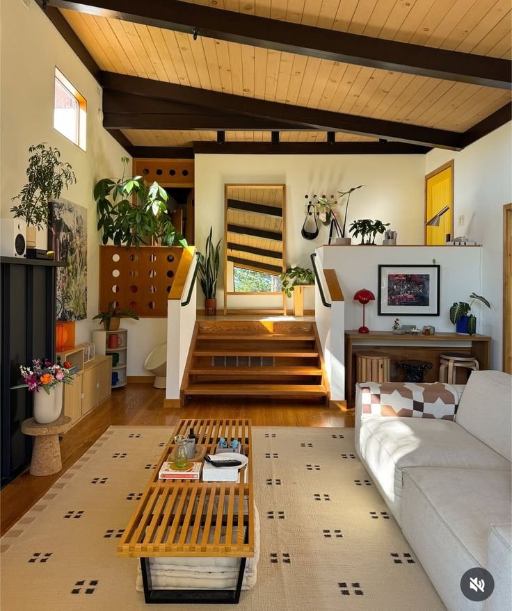 Living room with wooden stairs leading to a higher level, decorated with green plants, framed artwork, and a white sofa, with a cream-colored rug and a wooden coffee table in front.