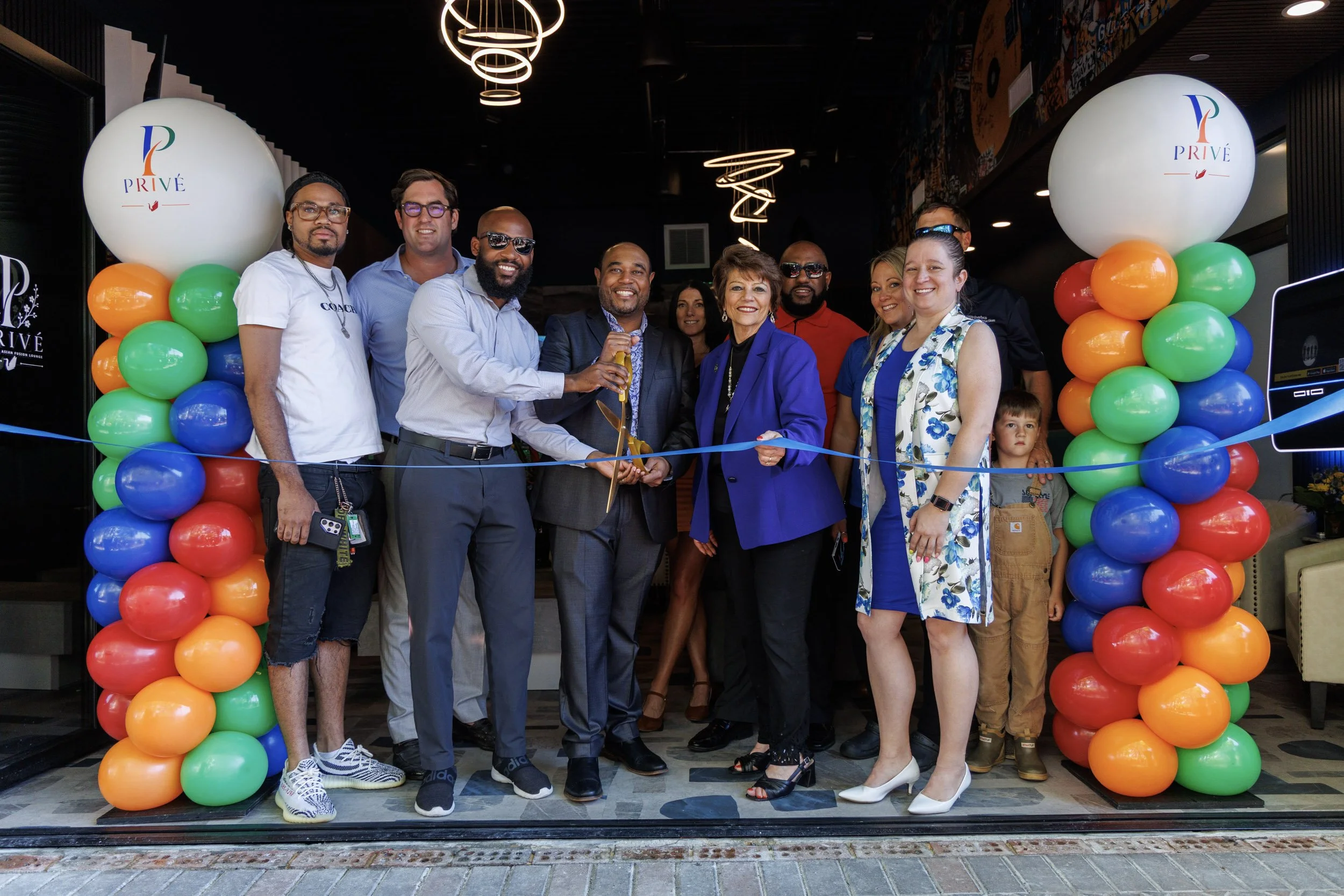 Group of people celebrating at an event, standing behind a ribbon, with rainbow-colored balloon arch and large white balloons with 'PRIVÉ' logo on either side.