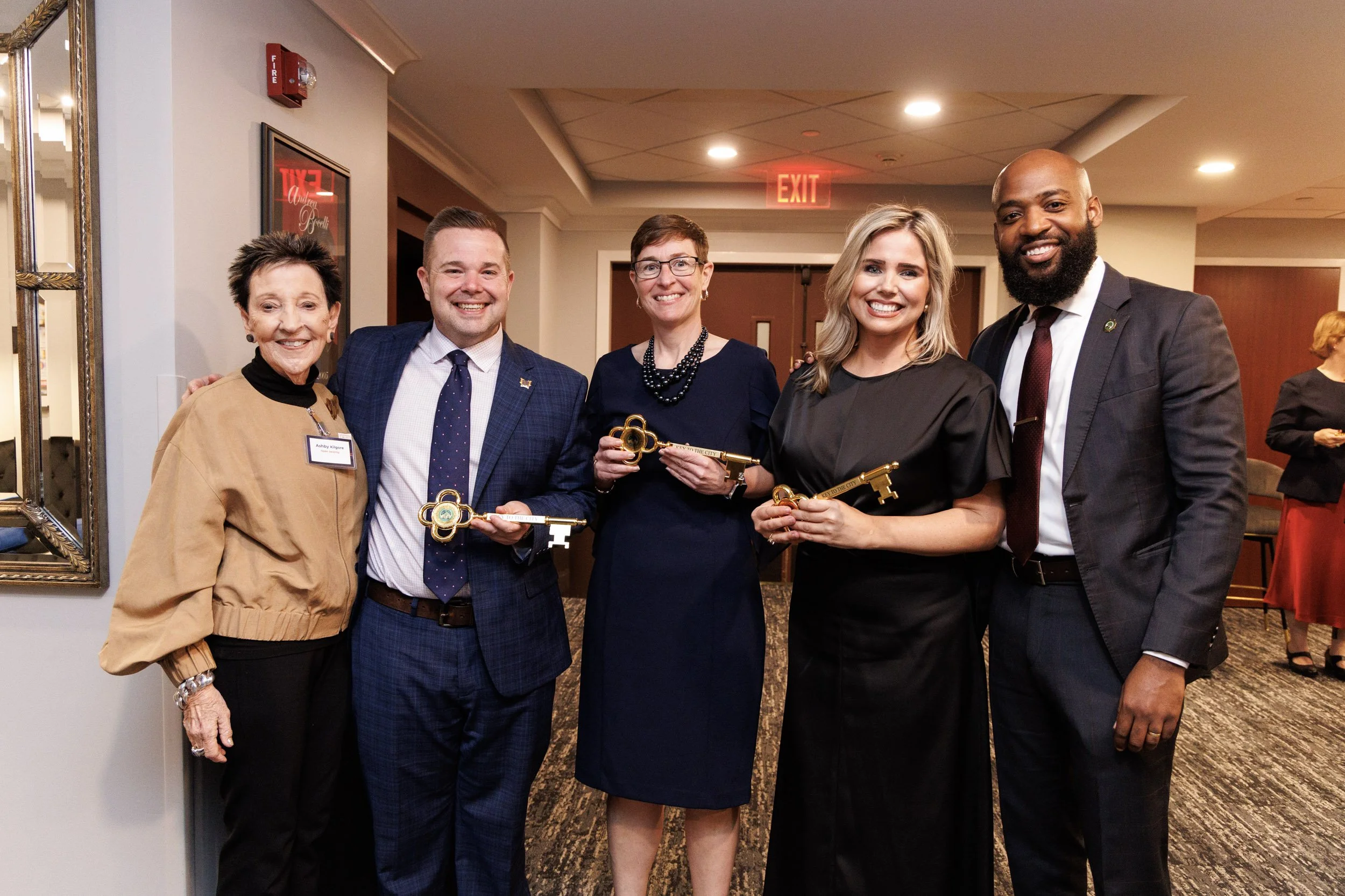A group of five diverse adults smiling and holding large antique-style keys at an indoor event, with a framed mirror on the wall and a lit exit sign in the background.