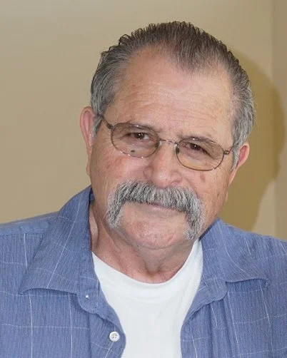 An older man with gray hair, a mustache, wearing glasses, a blue shirt over a white t-shirt, standing indoors against a neutral background.