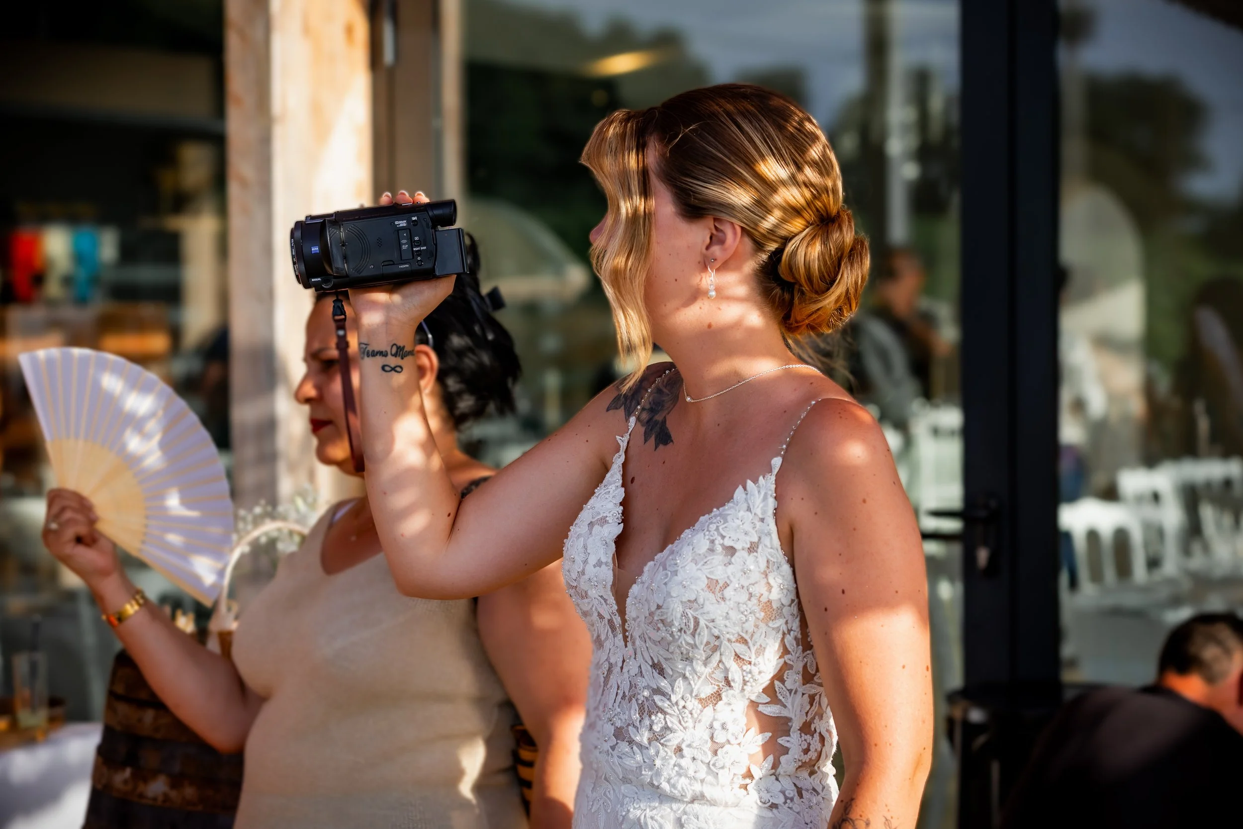Une femme en robe de mariage en train filmer avec une caméra Semper lors d'un événement en plein air.