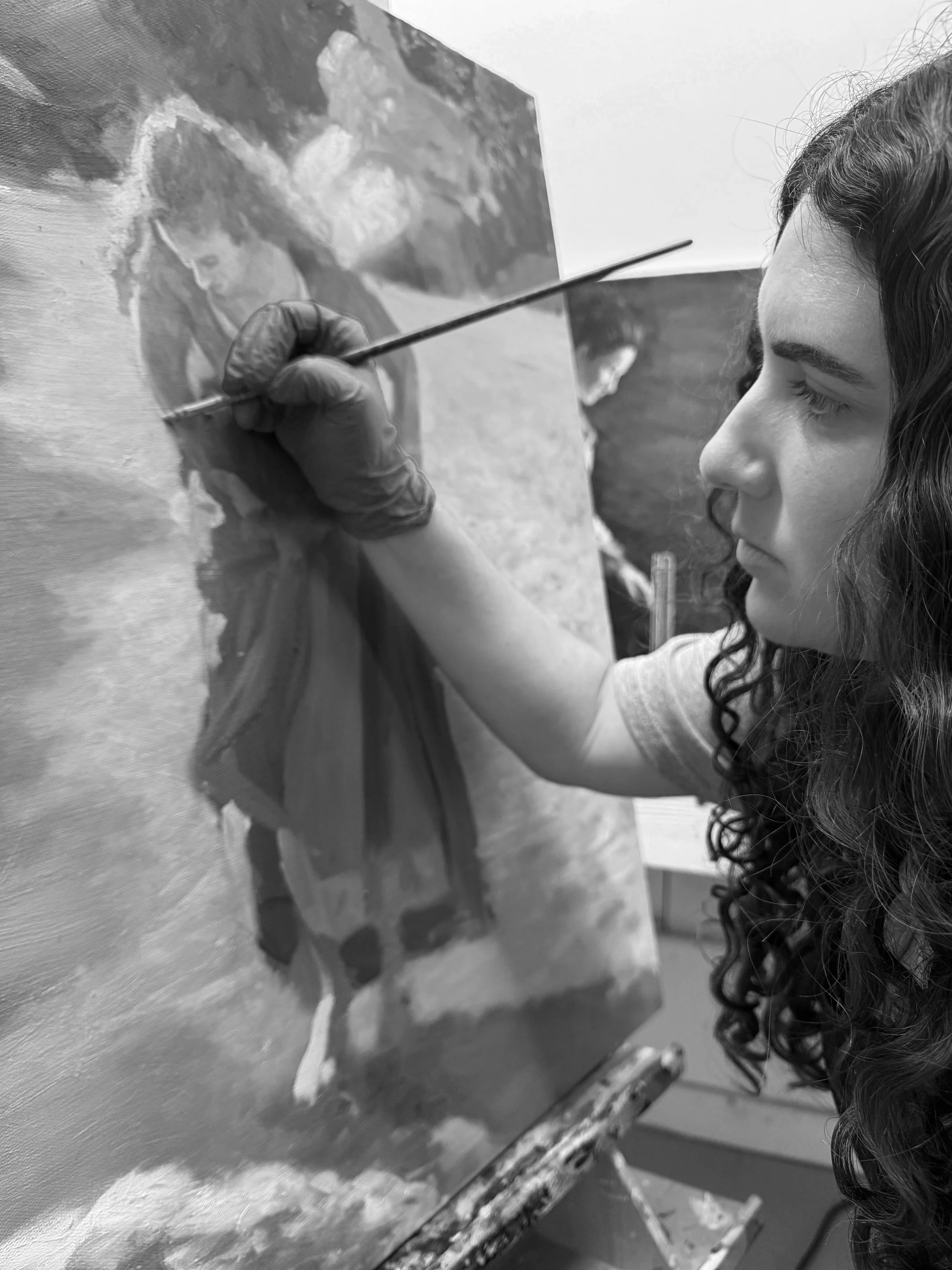 Black and white photo of a young woman with curly hair painting on a canvas. She is holding a paintbrush and focused on her artwork, which depicts another woman with similar features.