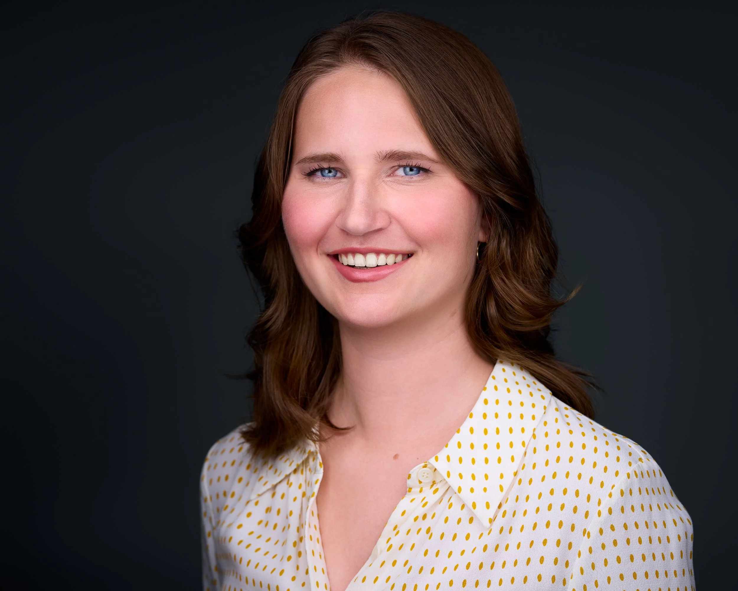 A young woman with shoulder-length brown hair, blue eyes, and a white blouse with yellow polka dots, smiling against a dark background.