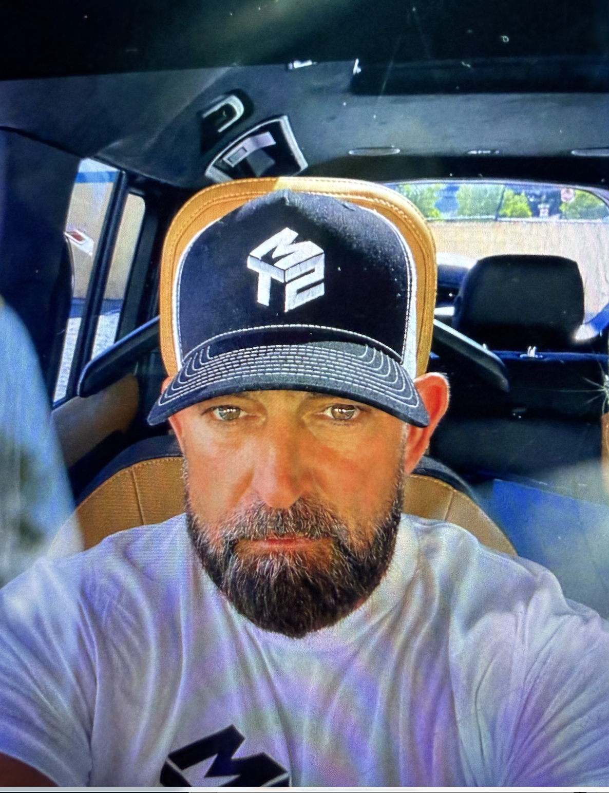A man with a beard, wearing a black and white trucker hat with a logo, is sitting in the back seat of a vehicle. The photo is taken from above, showing the man’s face, the hat, and part of the vehicle's interior, with a window showing trees and a blue sky in the background.