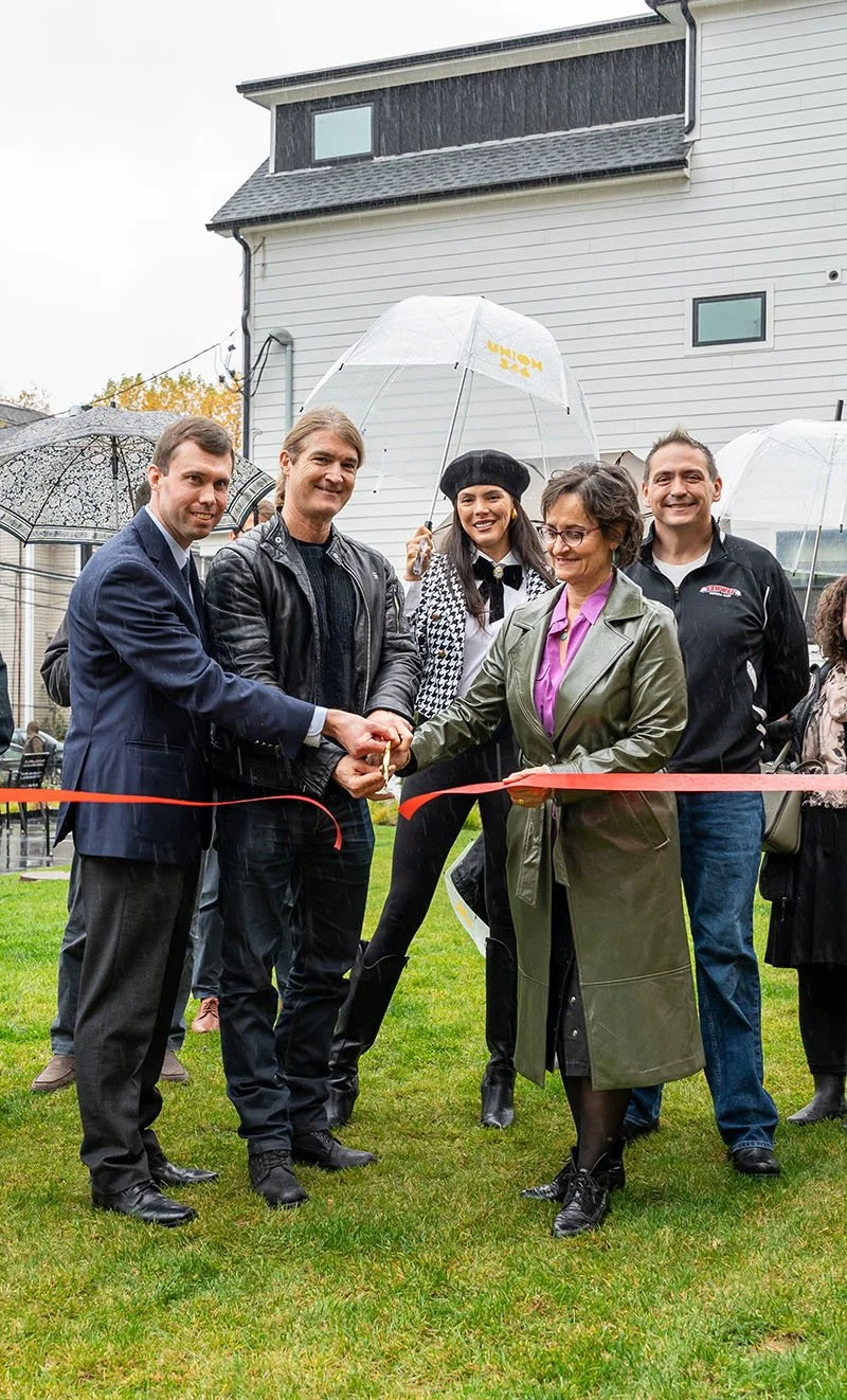 People standing outdoors during a ribbon-cutting ceremony on a rainy day, with some holding umbrellas and others wearing raincoats.