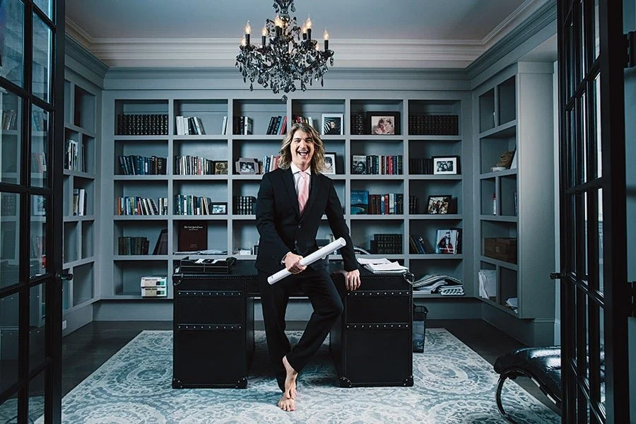 A woman in a business suit holding rolled-up papers, laughing happily in an elegant home office with bookshelves, a chandelier, and a desk.