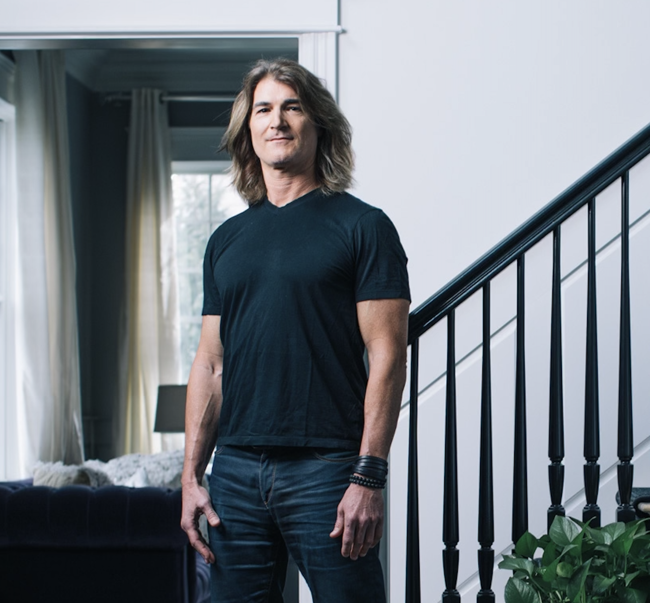 A man with long hair standing indoors next to a staircase, wearing a black t-shirt and dark jeans.