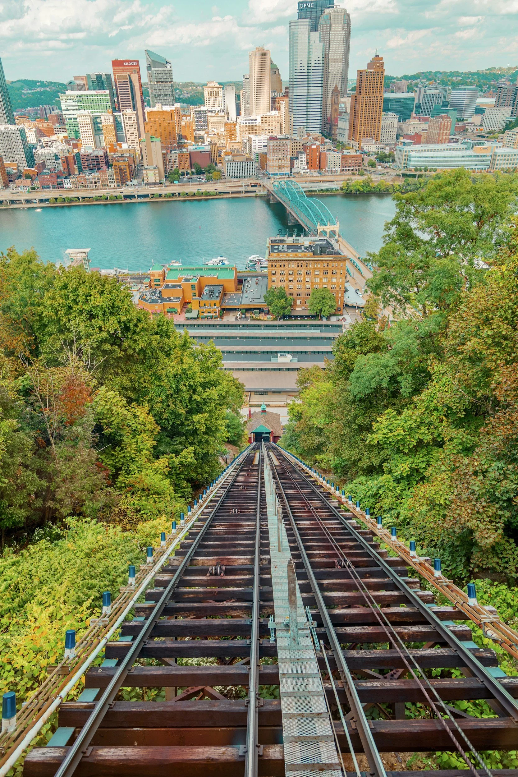 View from a funicular railway track looking down towards a city skyline with tall buildings, a river, and a bridge, surrounded by lush green trees.
