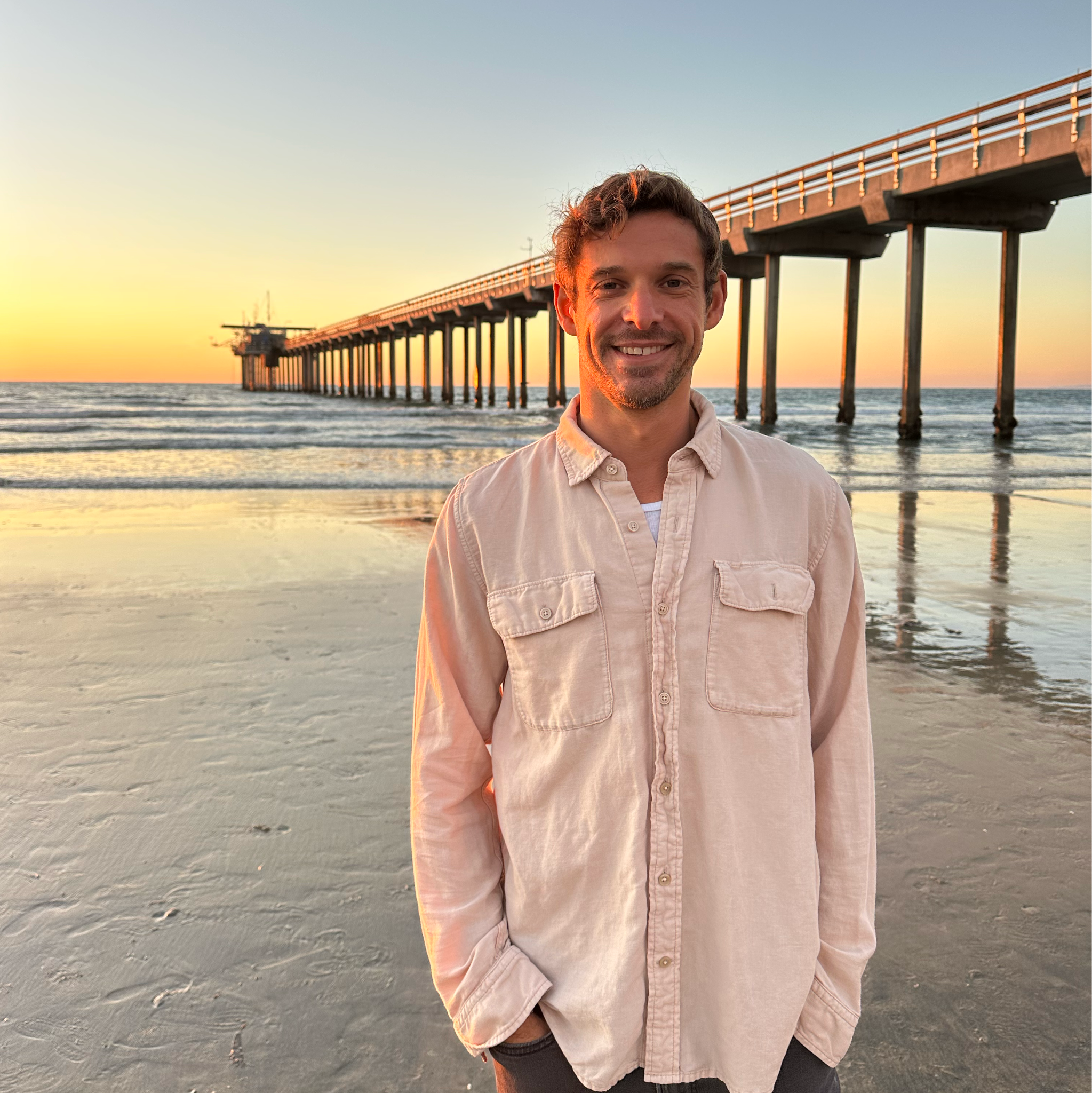 A man is standing on a beach with a pier extending into the ocean behind him at sunset.