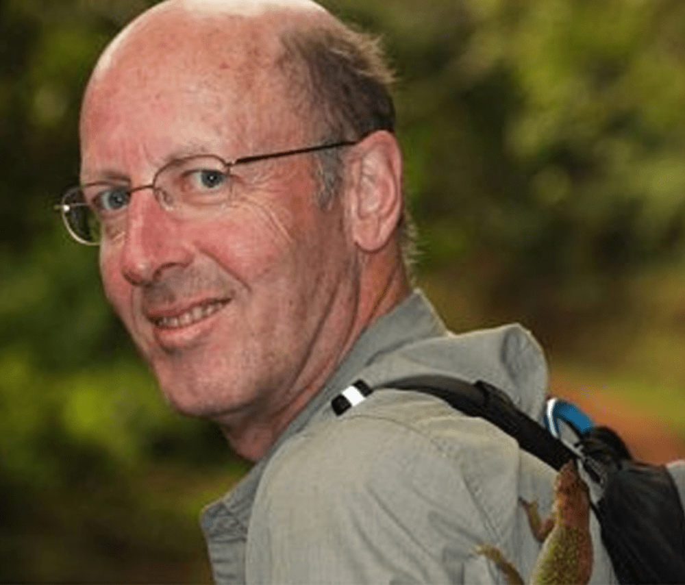 A man with glasses, a bald head, and a light beard outdoors with trees in the background, wearing a light-colored jacket and a backpack.
