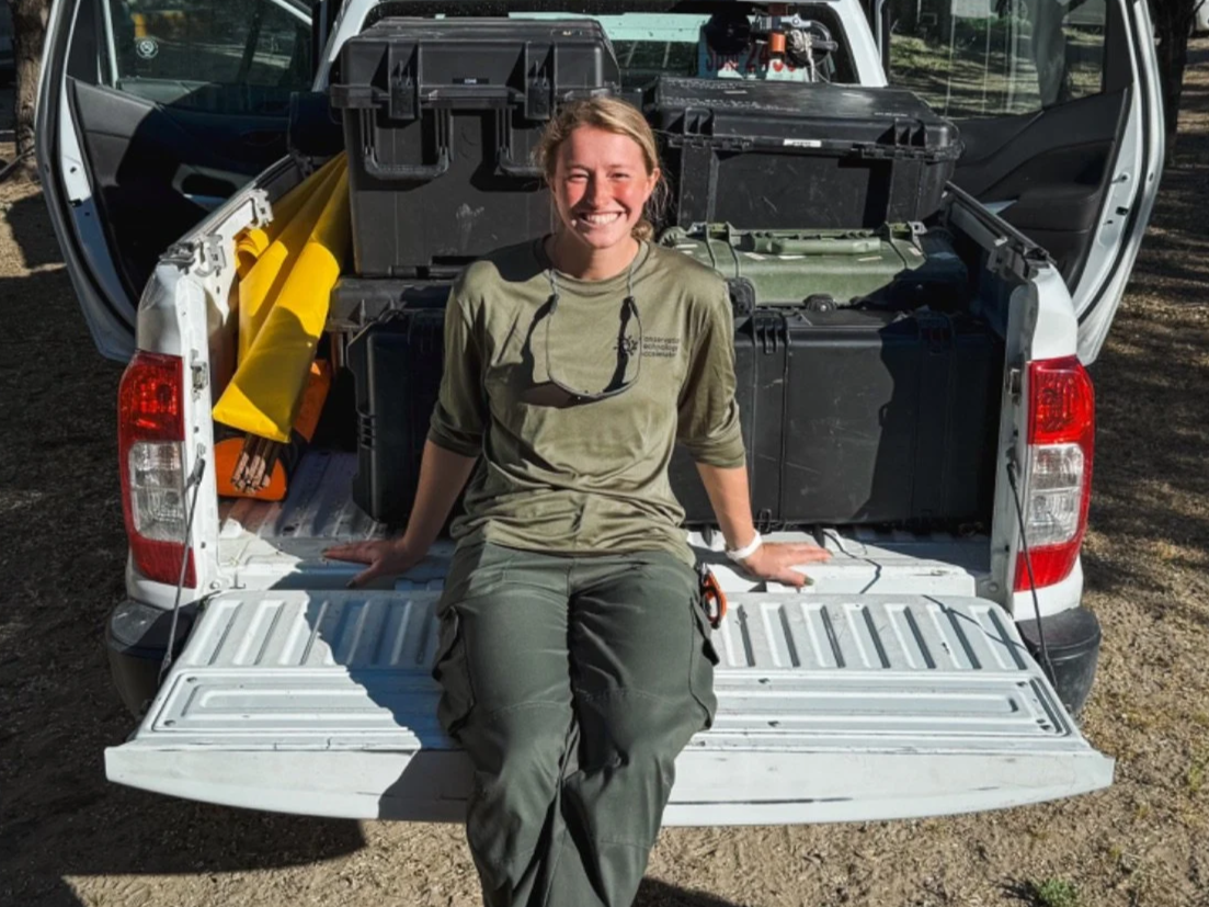 Lydia Morgantini sitting on the open tailgate of a pickup truck surrounded by large black and yellow toolboxes and equipment, smiling at the camera.
