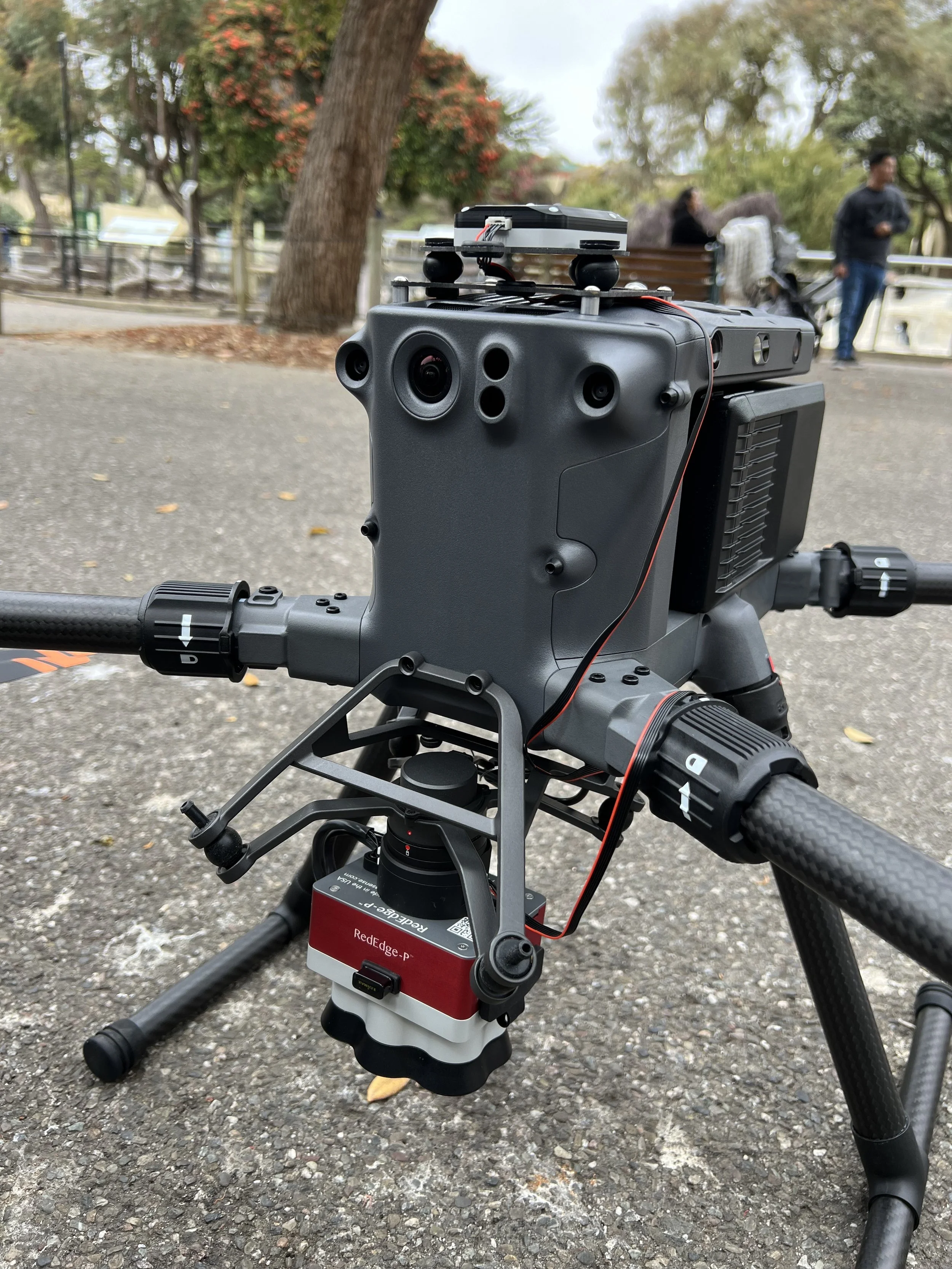 Close-up of a drone or robotic device mounted on a bicycle handlebar, with a park setting in the background, including trees and people walking.