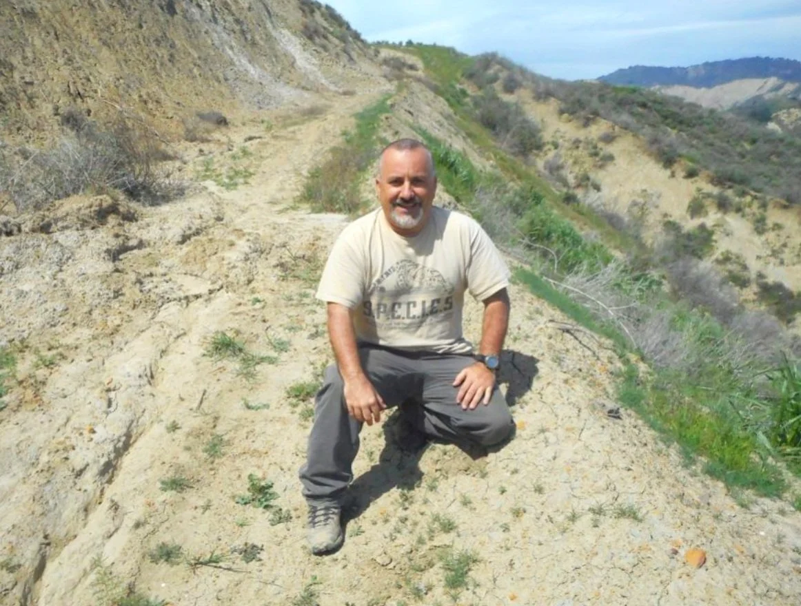 A man kneeling on a dirt trail along a hillside with sparse vegetation, with hills and a blue sky in the background.