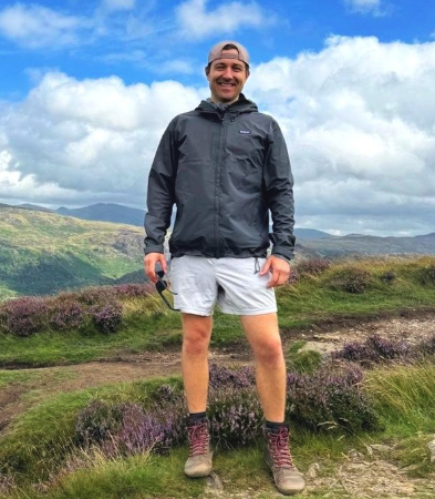 Sam Gray standing outdoors in a cloudy landscape, wearing a black jacket, white shorts, hiking boots, and a backward baseball cap.