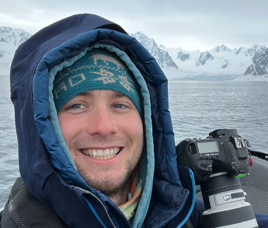Hunter Listwin smiling in a waterproof jacket and warm hat on a boat in icy waters with snow-covered mountains in the background, holding a camera.