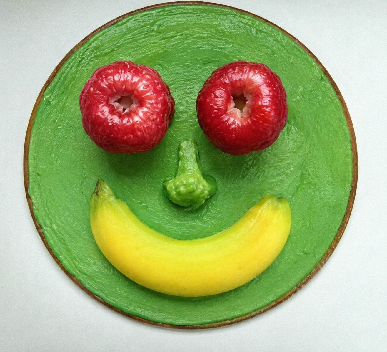 A green plate with a smiley face made of various fruits: two red apples for eyes, a green key lime for the nose, and a yellow banana for the mouth.