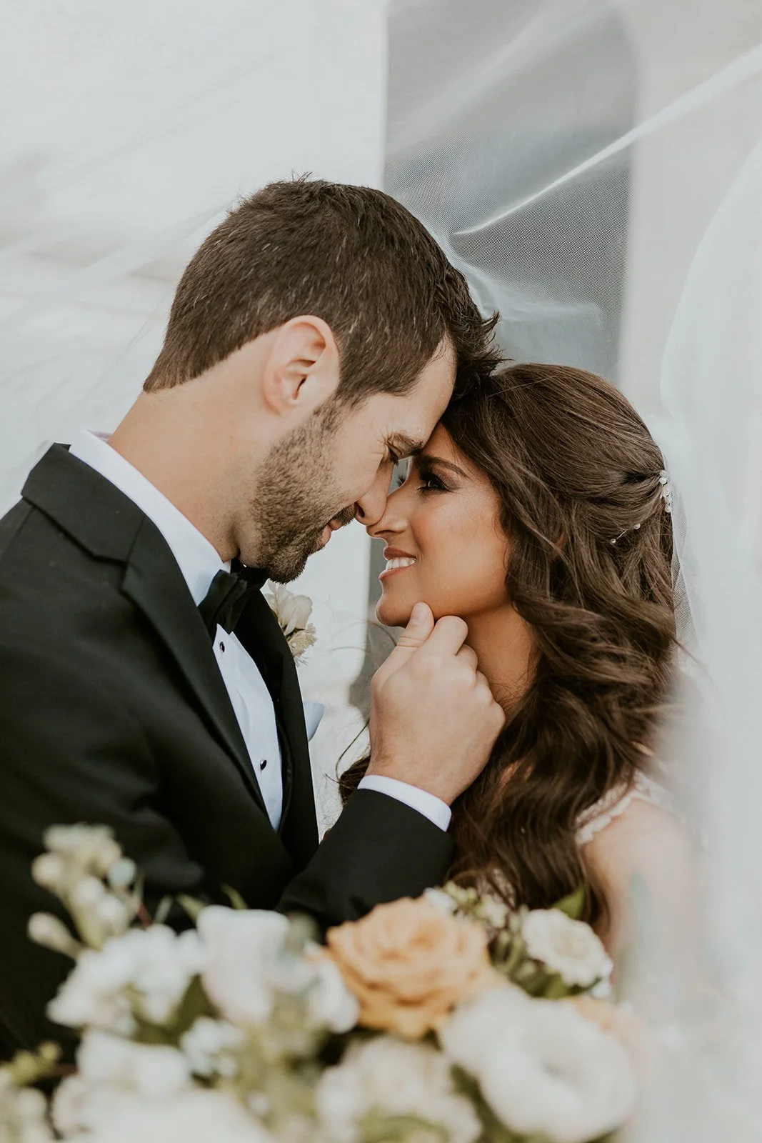 A bride and groom share a close, intimate moment, touching foreheads and smiling, with flowers in the foreground.