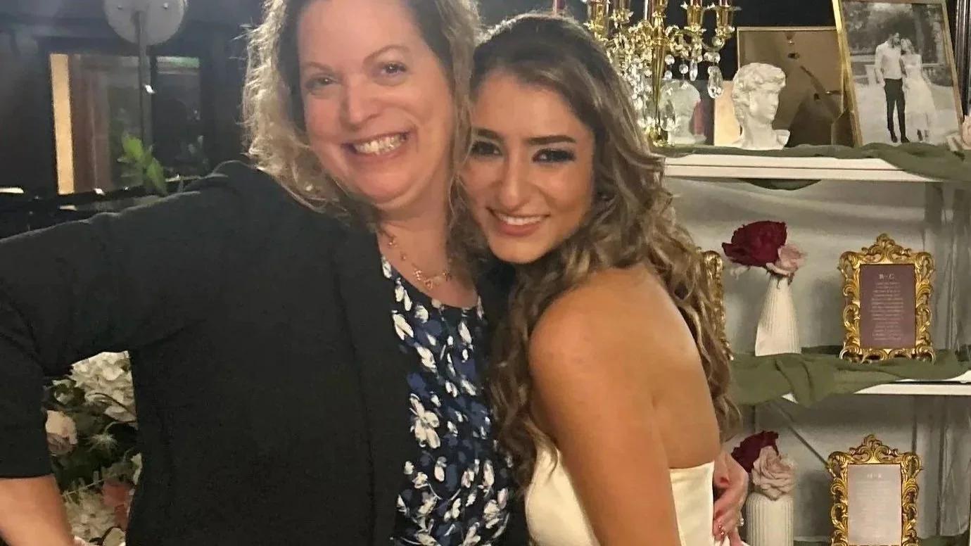 Two women smiling and posing for a photo at a celebration or event. One woman has curly blonde hair and is wearing a black blazer, while the other has long wavy hair and is wearing a strapless dress. In the background, there are decorative items including framed photos, flowers, a candlestick, and bust sculptures.