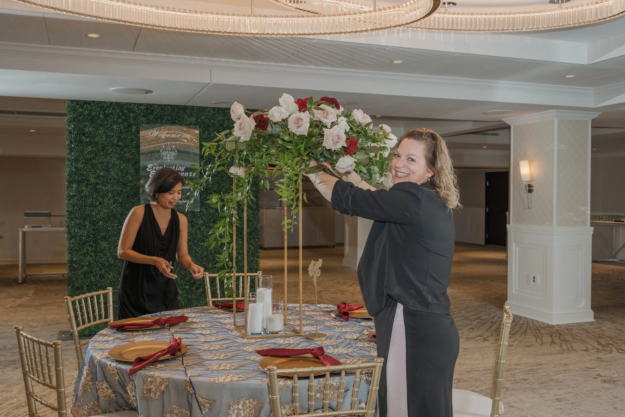 A woman with curly blonde hair in a black blazer smiling as she puts a bouquet of white and red flowers on a decorated table in a banquet hall. Another woman with short dark hair in a black dress is arranging items on the table. The table has a blue and gold tablecloth, gold plates with maroon napkins, and white candles. The background features a green hedge wall with a