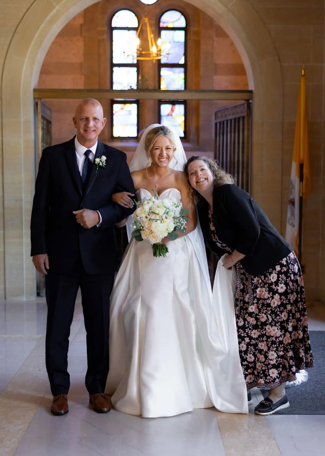 A bride in a wedding gown holding a bouquet, standing between a man in a suit and a woman in a floral dress, inside a church with stained glass windows and a chandelier.