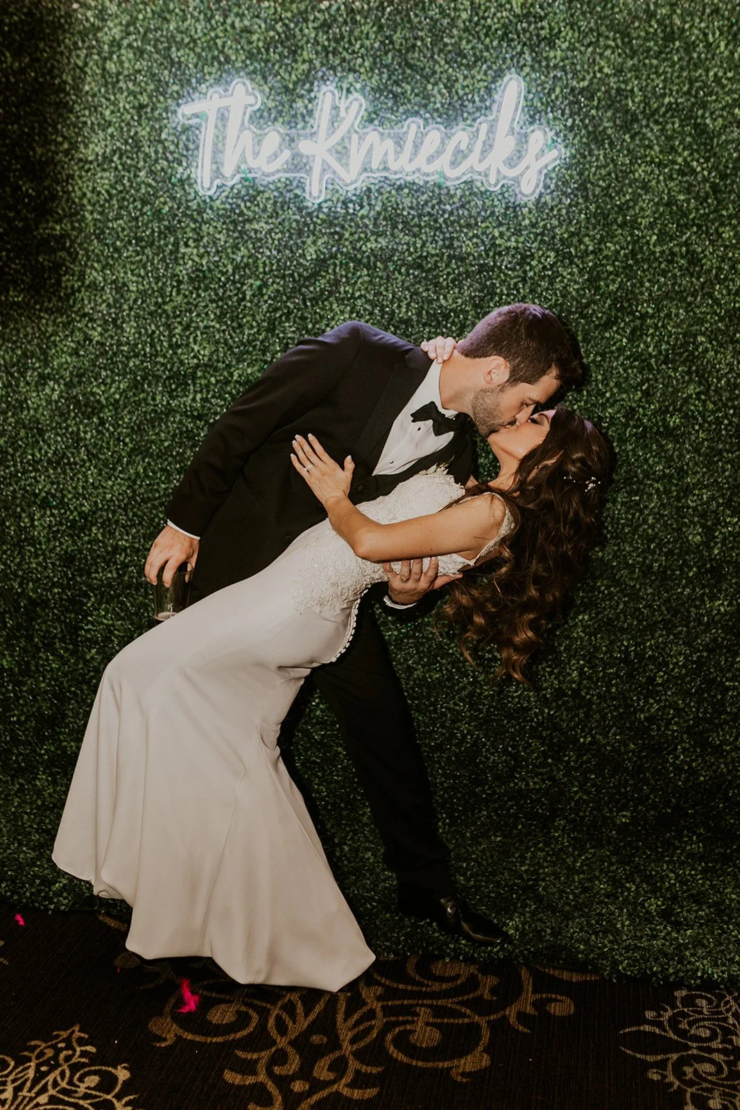 A couple dressed in wedding attire sharing a kiss while being dipped by the groom in front of a green hedge wall with neon sign reading 'The Knacks'.