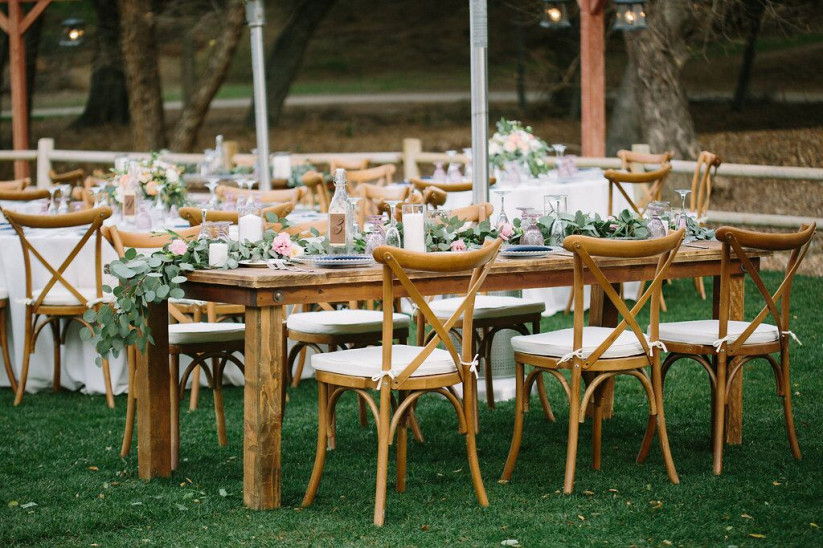 Outdoor wedding reception setup with wooden tables and chairs, decorated with white and pink floral centerpieces and greenery.