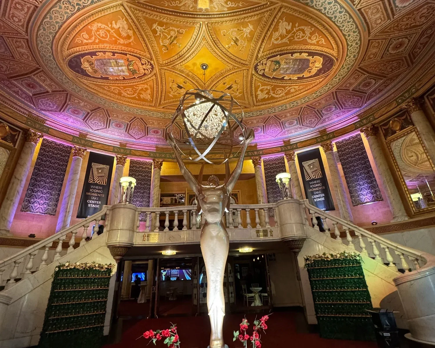 Elegant hotel lobby featuring a sculpture of a woman holding a globe-like structure above her head, ornate ceiling with decorative paintings, banners with text, and grand staircase.