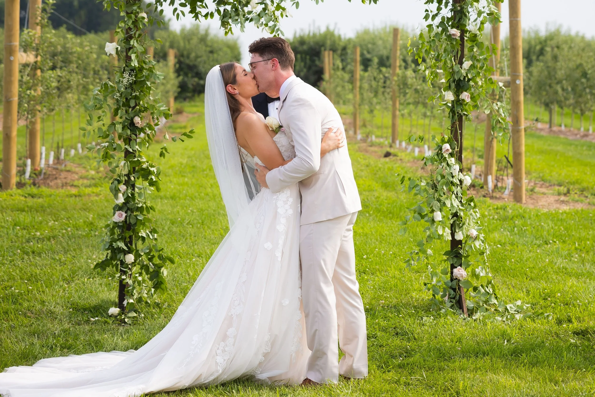 A bride and groom kiss under a floral arch on their wedding day in a lush outdoor setting.