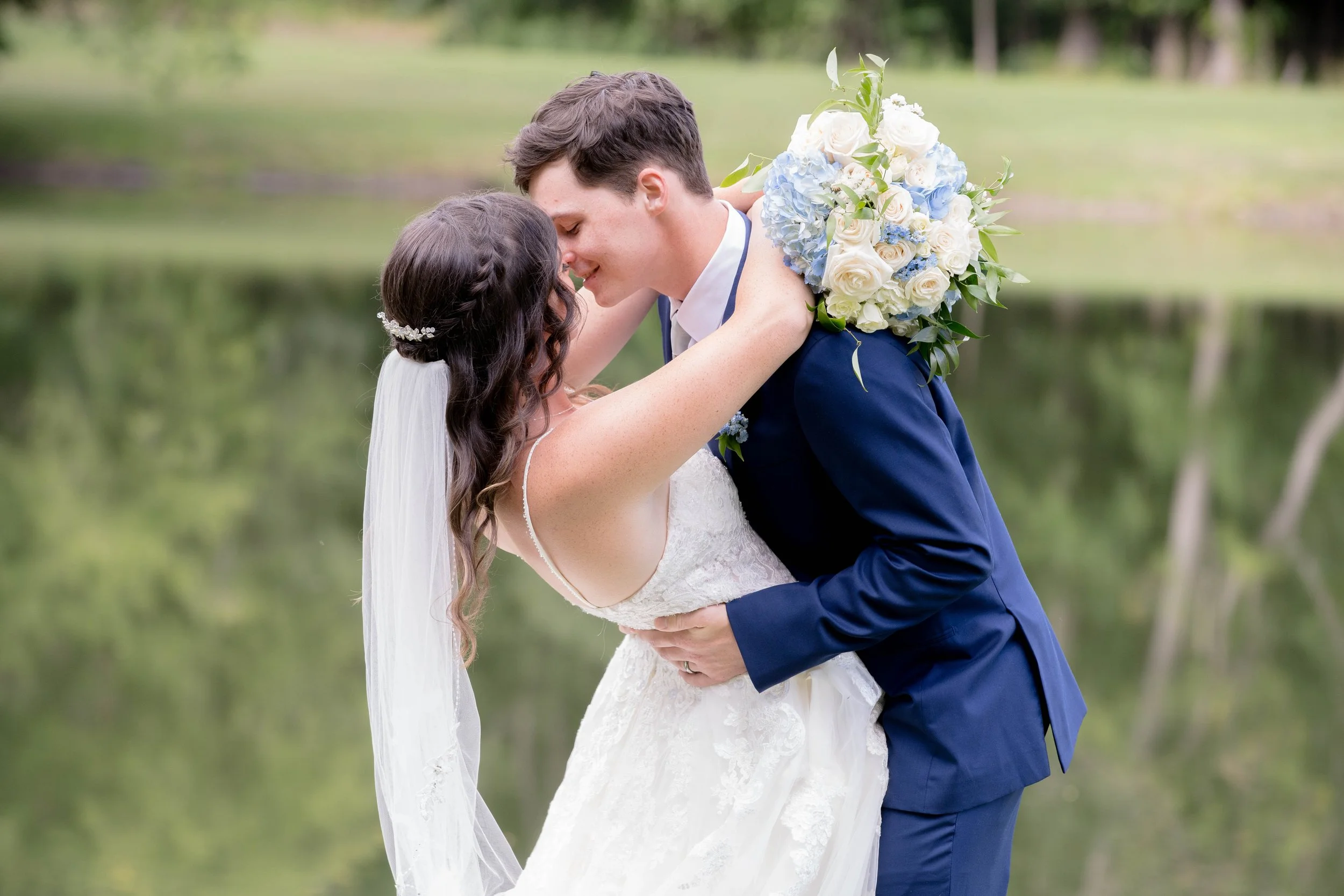 A newlywed couple shares a romantic moment outdoors near a lake, with the groom holding the bride, who wears a white wedding dress and veil, and the groom in a blue suit holding a bouquet of white and blue flowers.