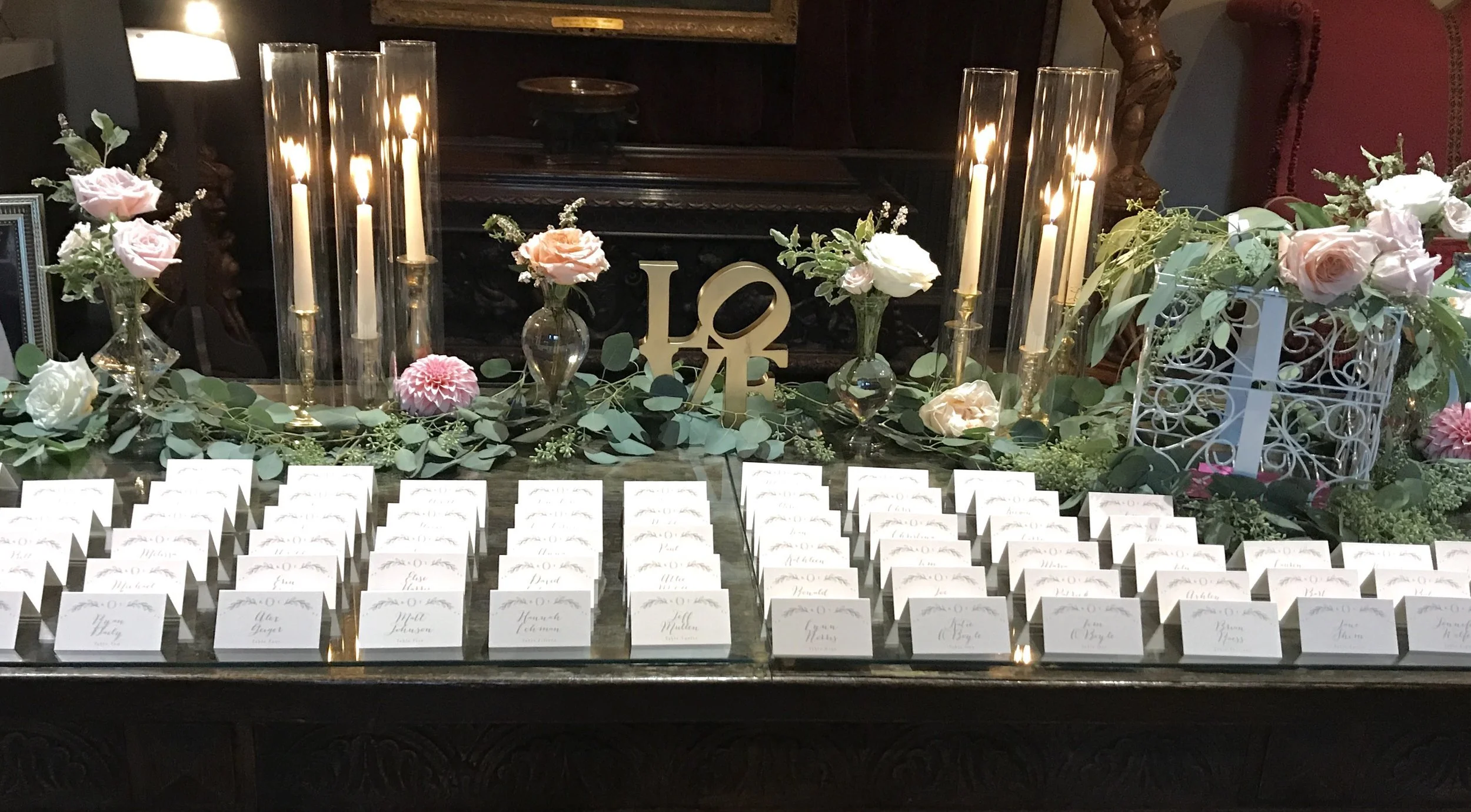 Table decorated with pink and white roses, greenery, lit candles, and a gold "LOVE" sign, with place cards arranged in front for an event.