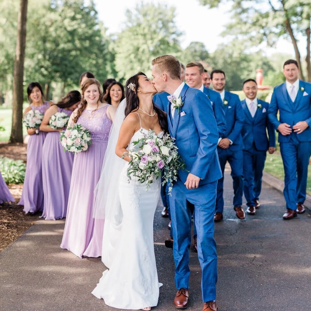 A bride and groom kiss in the center while wedding party members smile and watch in the background outdoors.