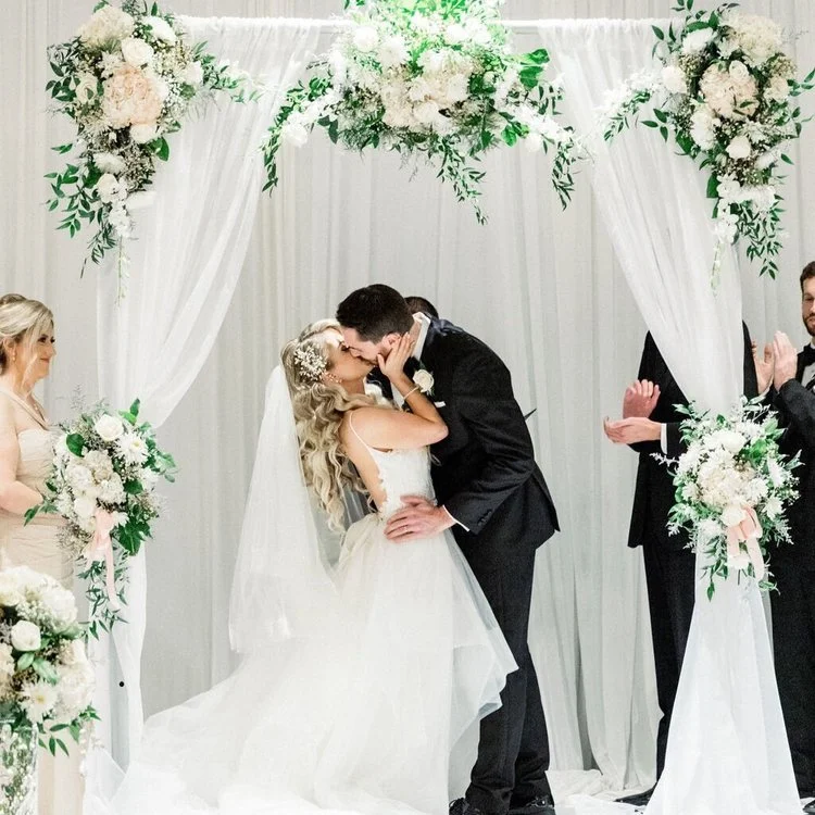 A bride and groom share a kiss during their wedding ceremony under a floral arch, with bridesmaids and groomsmen clapping nearby.