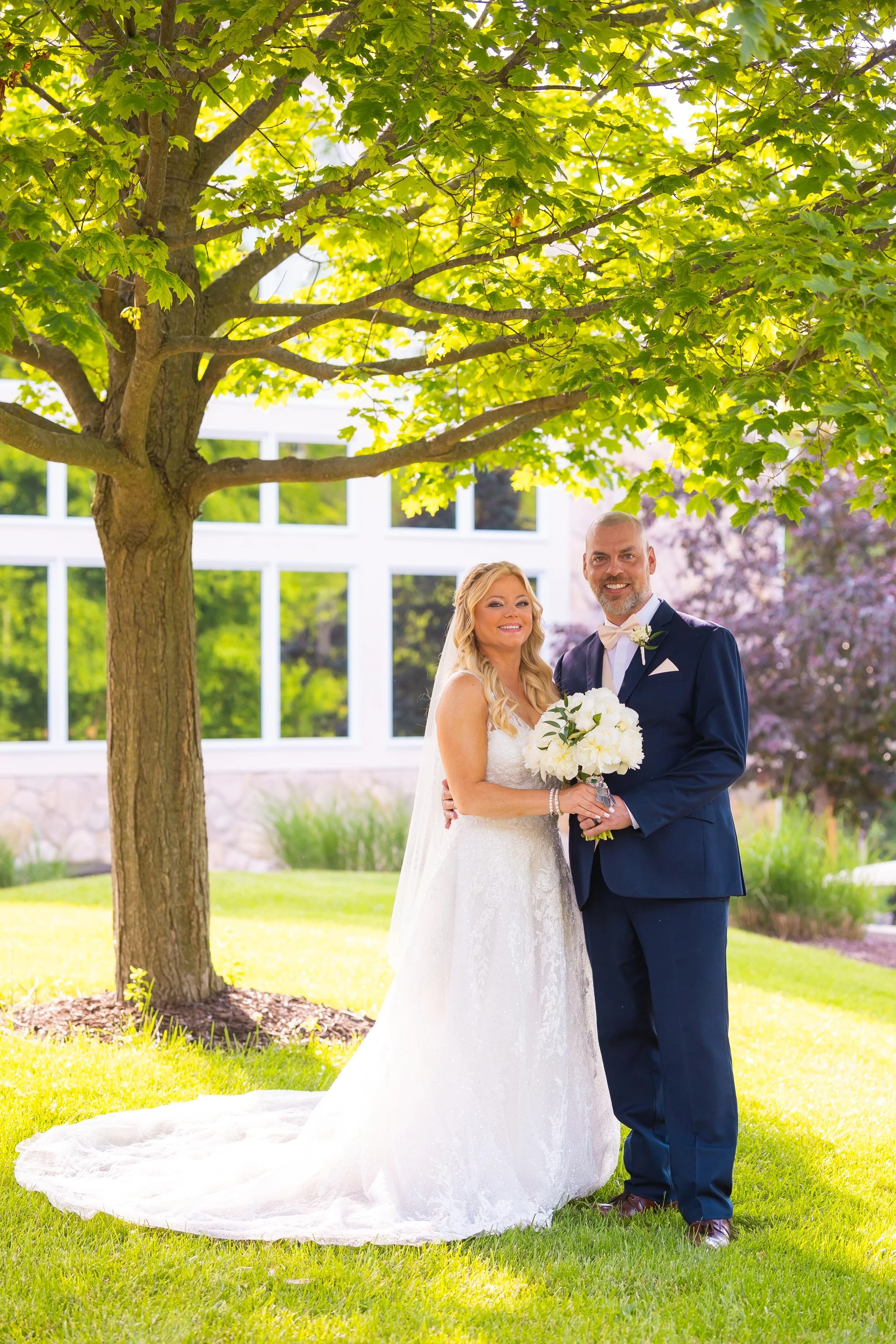 A bride and groom standing outdoors under a large green tree, smiling and posing for a wedding photo. The bride is holding a bouquet of white flowers and wearing a white wedding gown, while the groom is dressed in a navy blue suit with a white shirt and a light-colored bow tie.