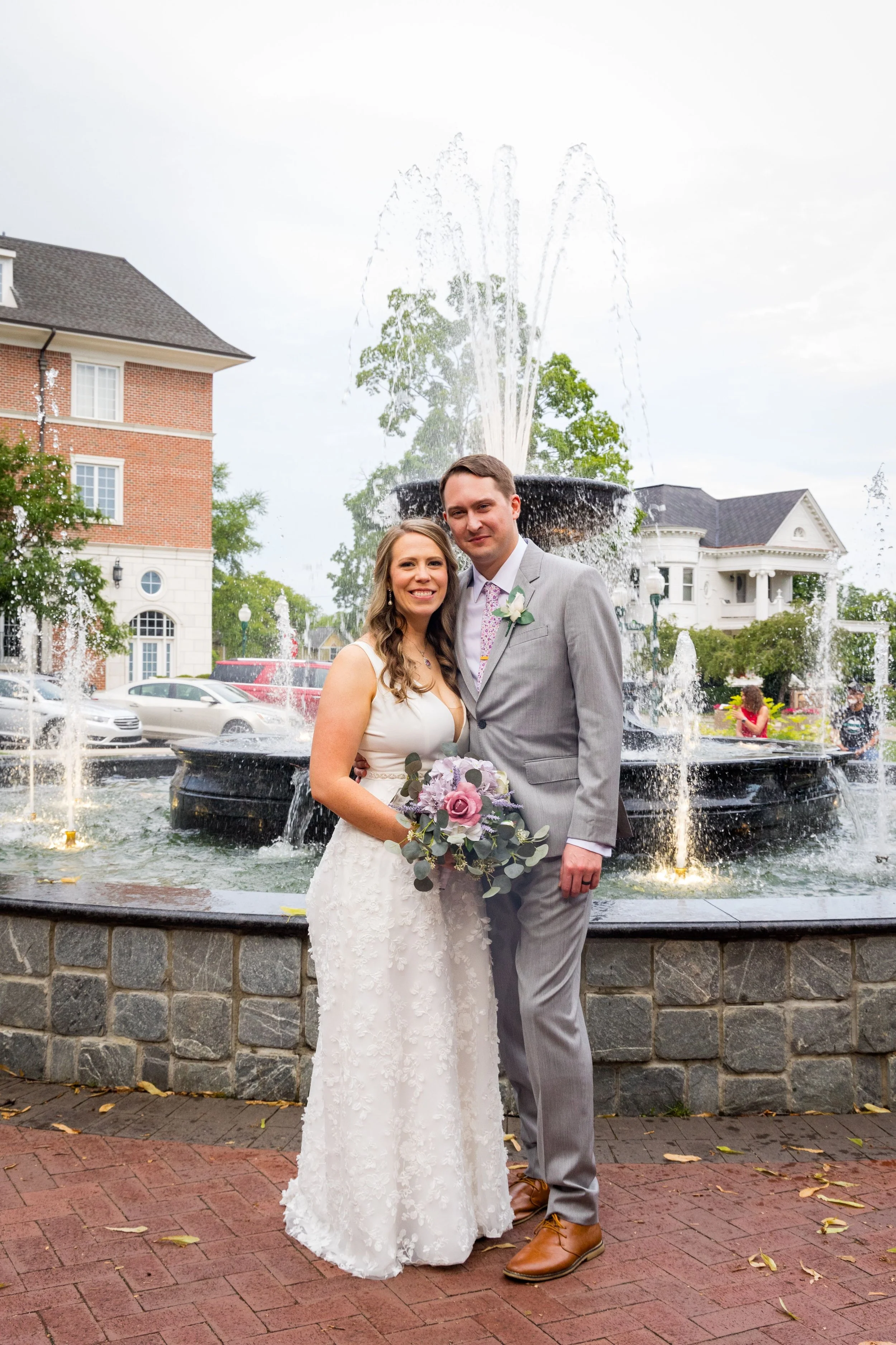 A bride and groom posing in front of a fountain during their wedding day. The bride wears a white lace wedding dress and holds a bouquet, while the groom wears a gray suit with a pink tie and a boutonniere. There are historic buildings and trees in the background.