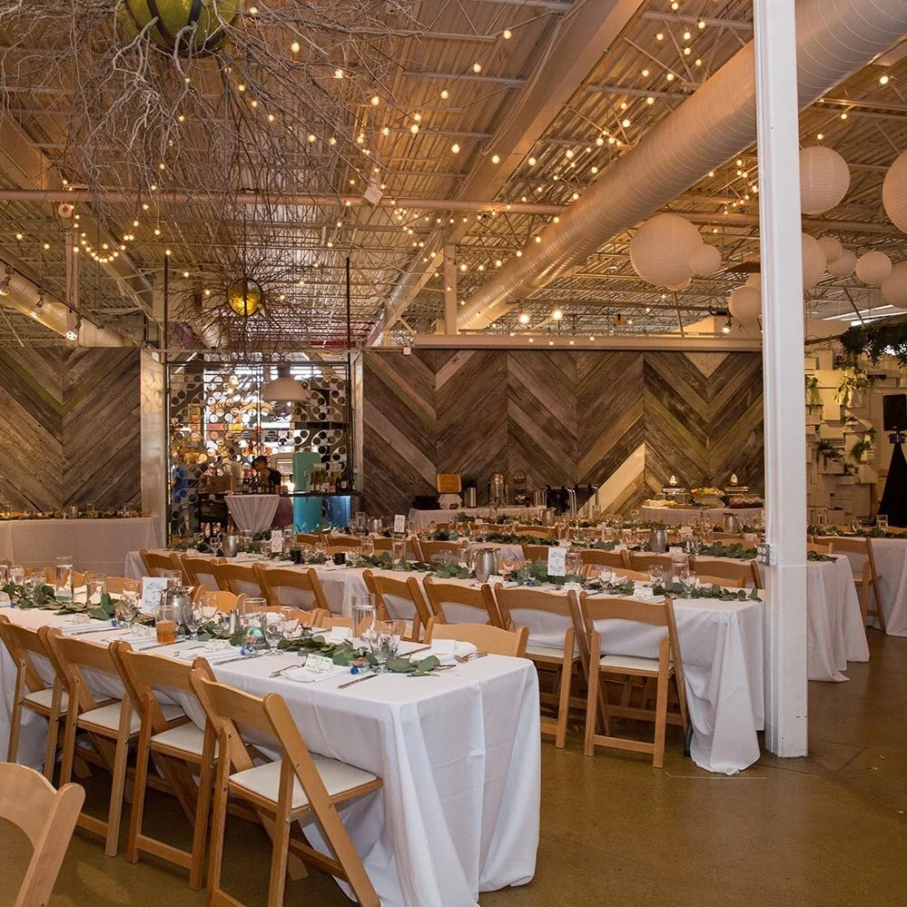 Indoor wedding reception setup with long tables covered in white tablecloths, decorated with greenery and place settings, surrounded by wooden chairs. The ceiling is decorated with string lights and paper lanterns, and a wooden accent wall is visible in the background.