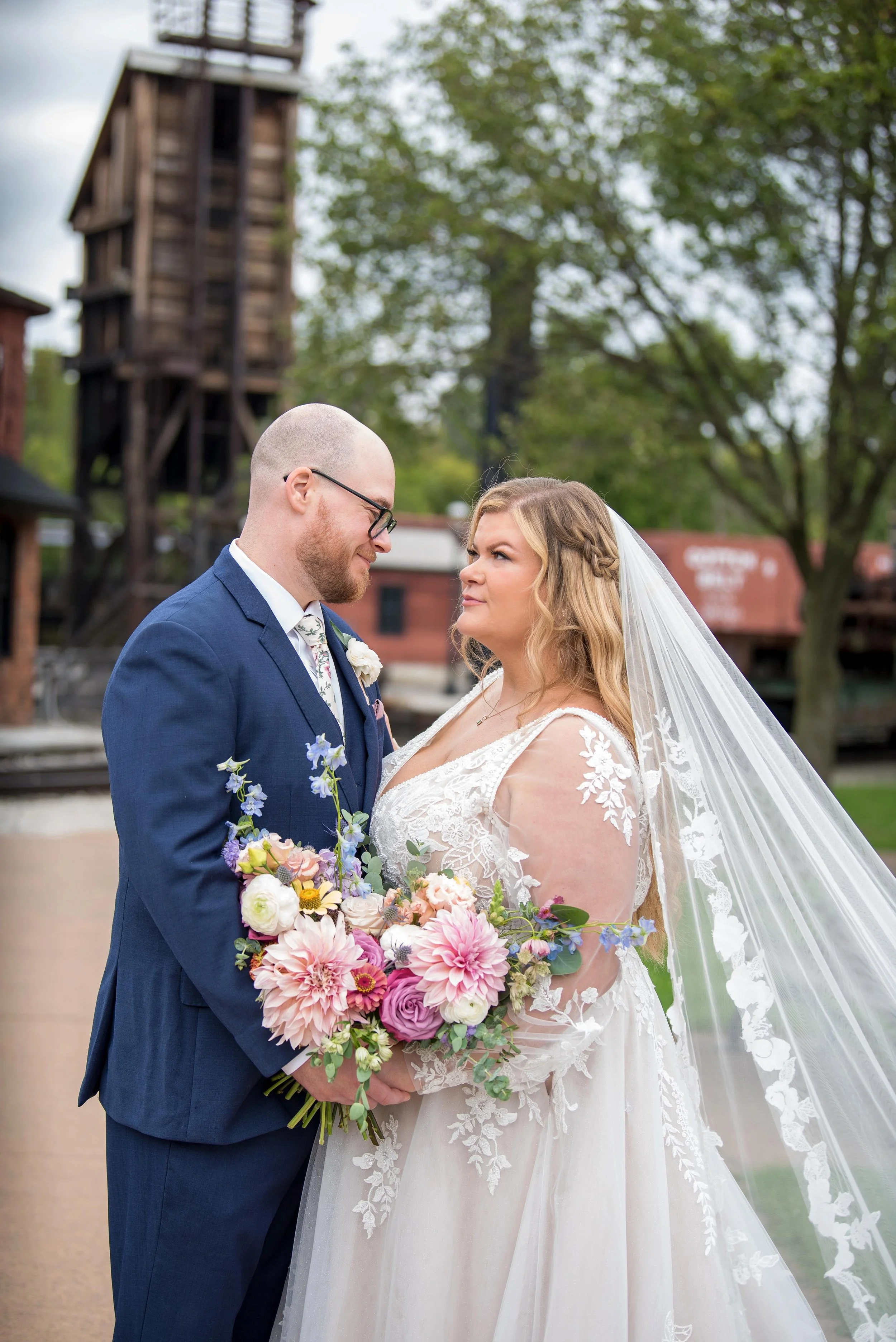 A bride and groom stand close together outdoors on their wedding day, with the groom wearing a blue suit and the bride in a white lace wedding dress and veil, holding a colorful bouquet of flowers.