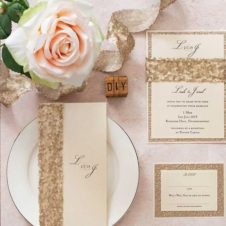 Wedding invitations with gold sequined accents, a white plate, a pink and white rose, and a wooden 'DIY' block on a soft pink background.