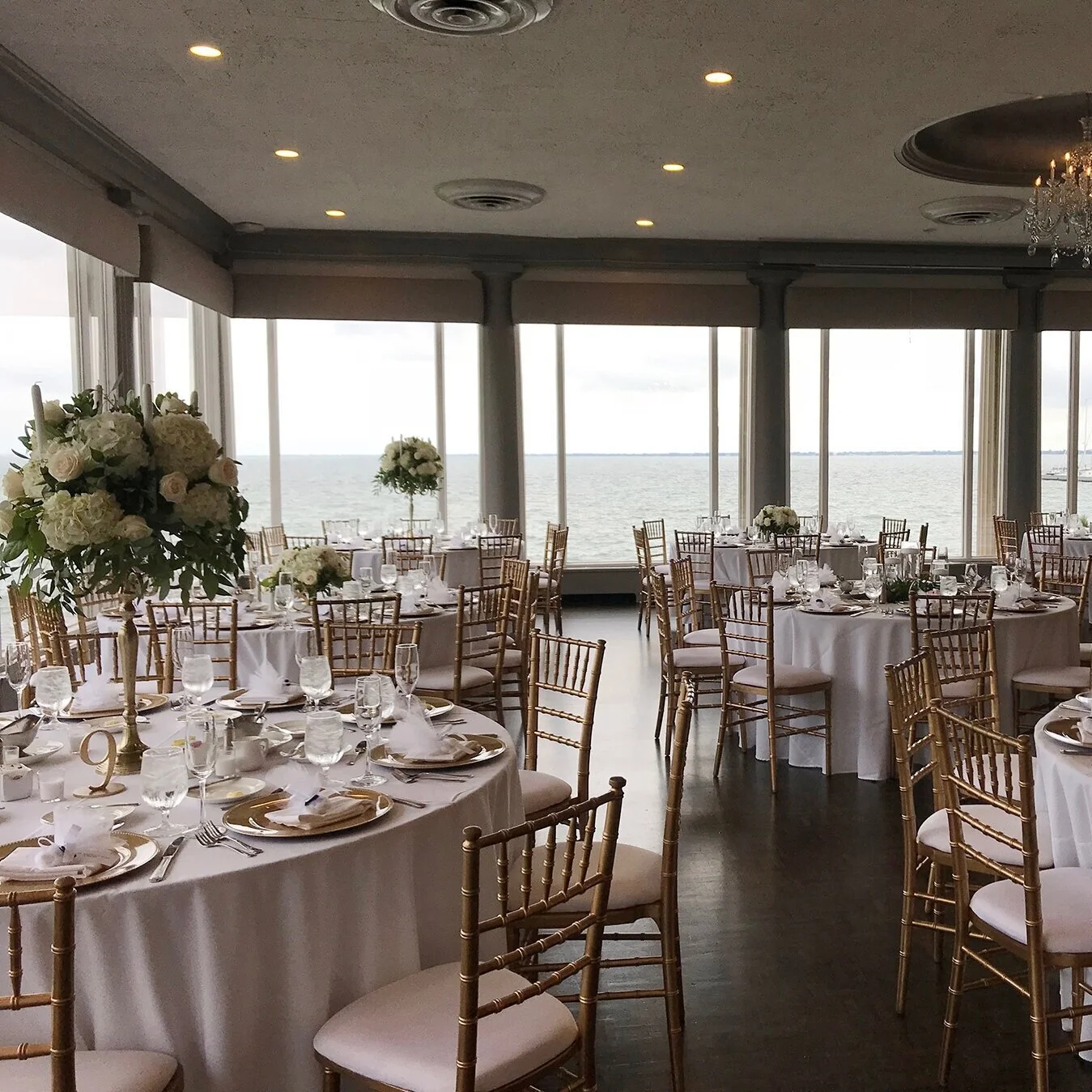 Elegant banquet hall with round tables adorned with white tablecloths, floral centerpieces, and gold chairs, overlooking a waterfront view through large windows.