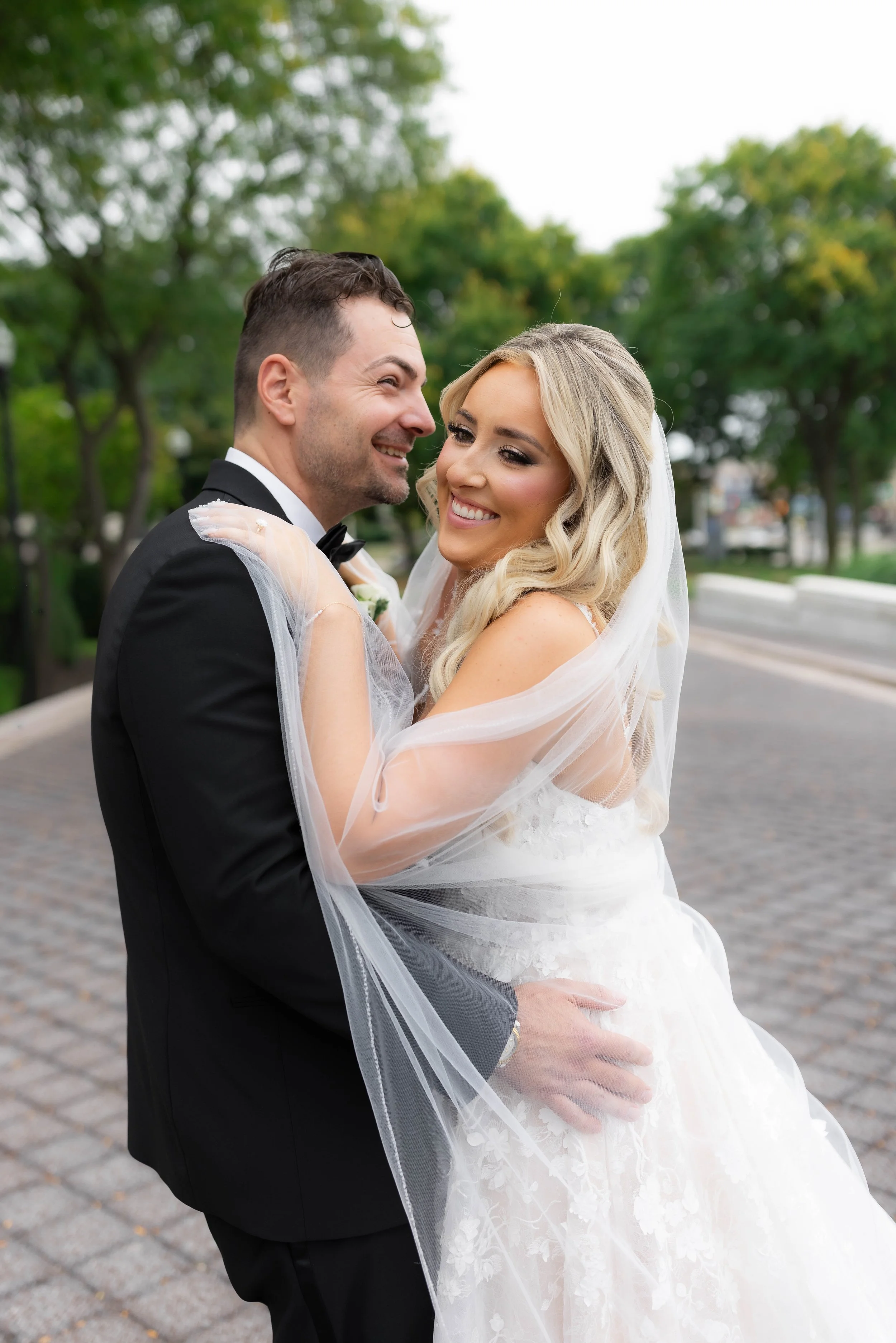 A bride and groom smiling and embracing outdoors, with trees and a pathway in the background.