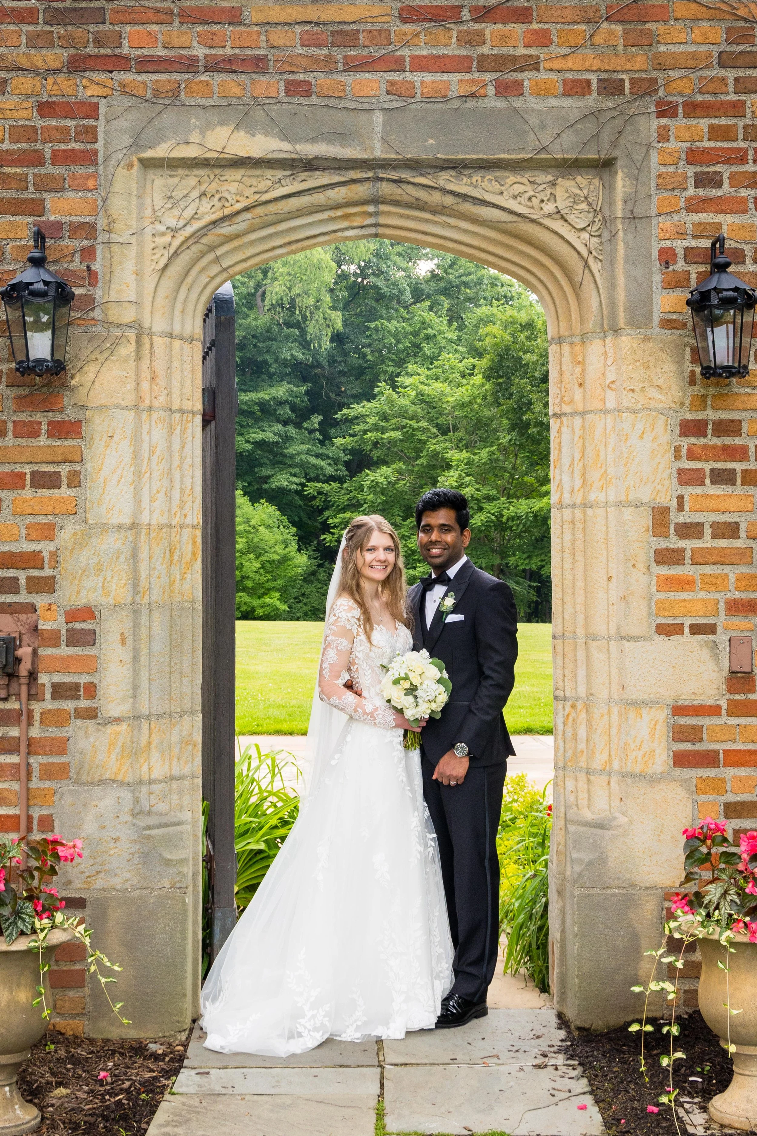 A bride and groom standing together under an ornate stone archway with greenery in the background. The bride is in a white wedding dress holding a bouquet, and the groom is in a black tuxedo with a bow tie.