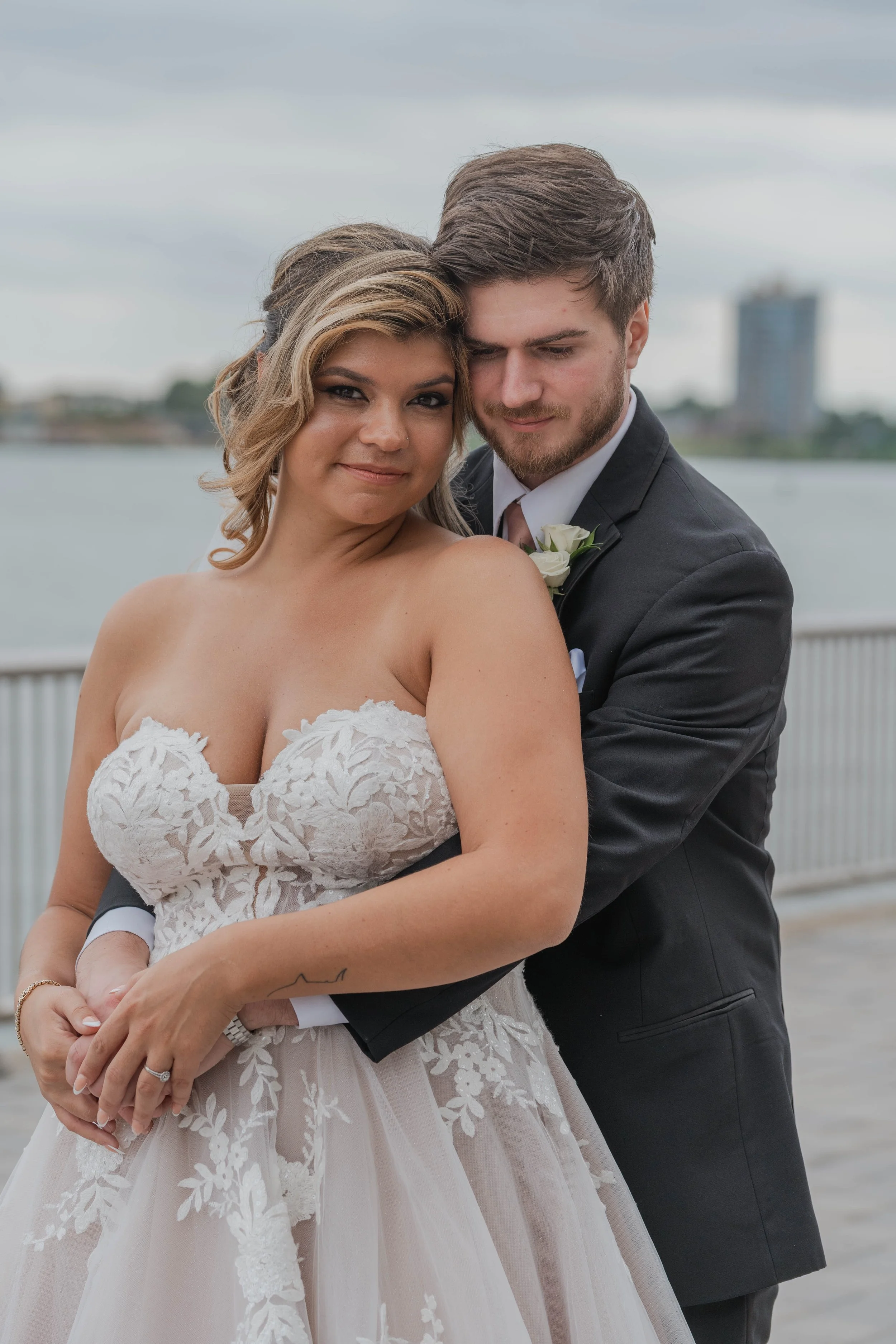 A bride and groom are embracing outdoors, with water and city buildings in the background. The bride is wearing a lace wedding dress and the groom is in a dark suit with a white shirt and a boutonniere.