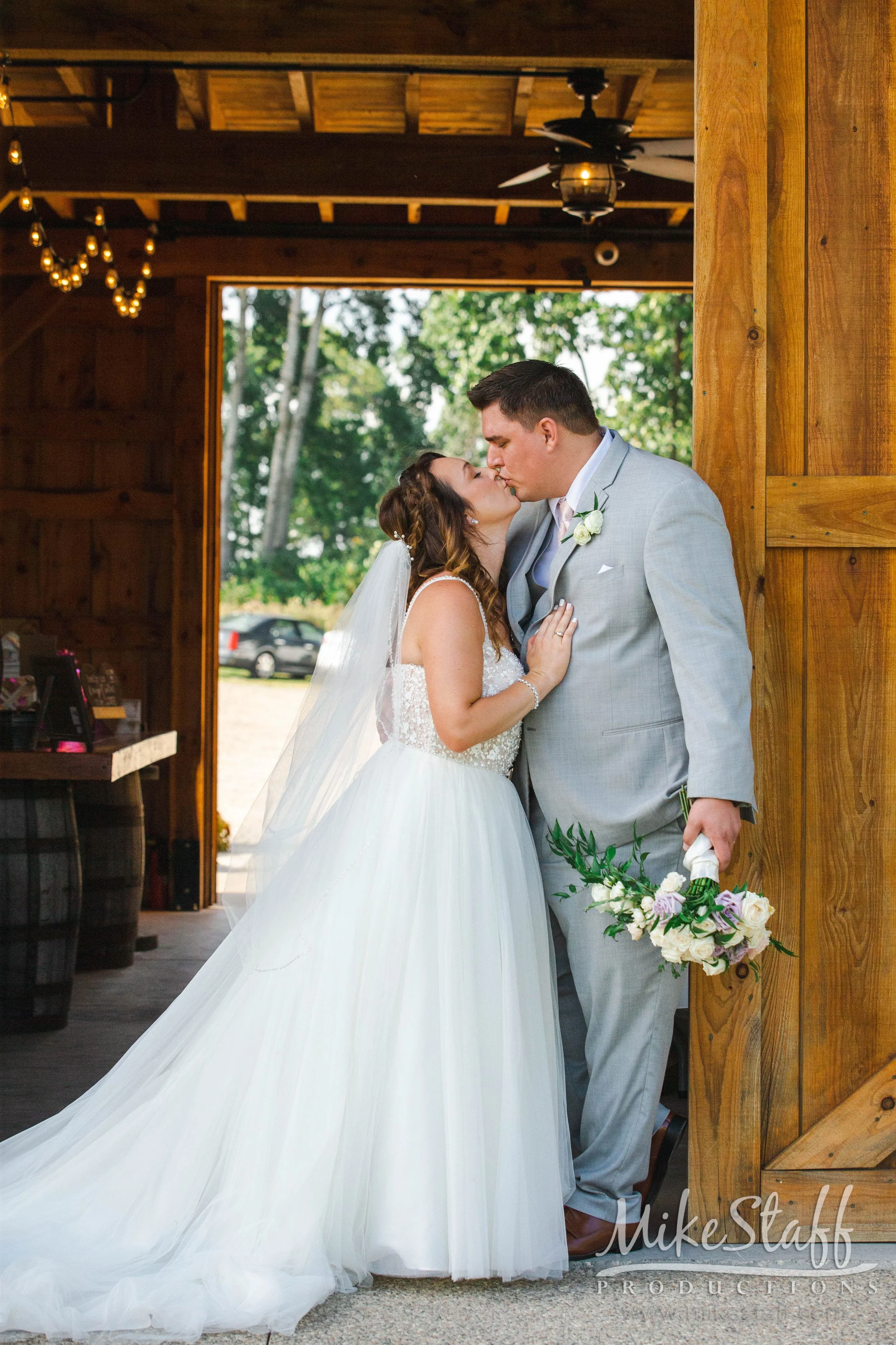 A bride and groom share a kiss inside a rustic wooden building with large open doorway and string lights hanging from the ceiling. The bride wears a white strapless wedding gown with a long tulle train, and the groom wears a light gray suit with a white boutonniere, holding a bouquet of white and purple flowers.