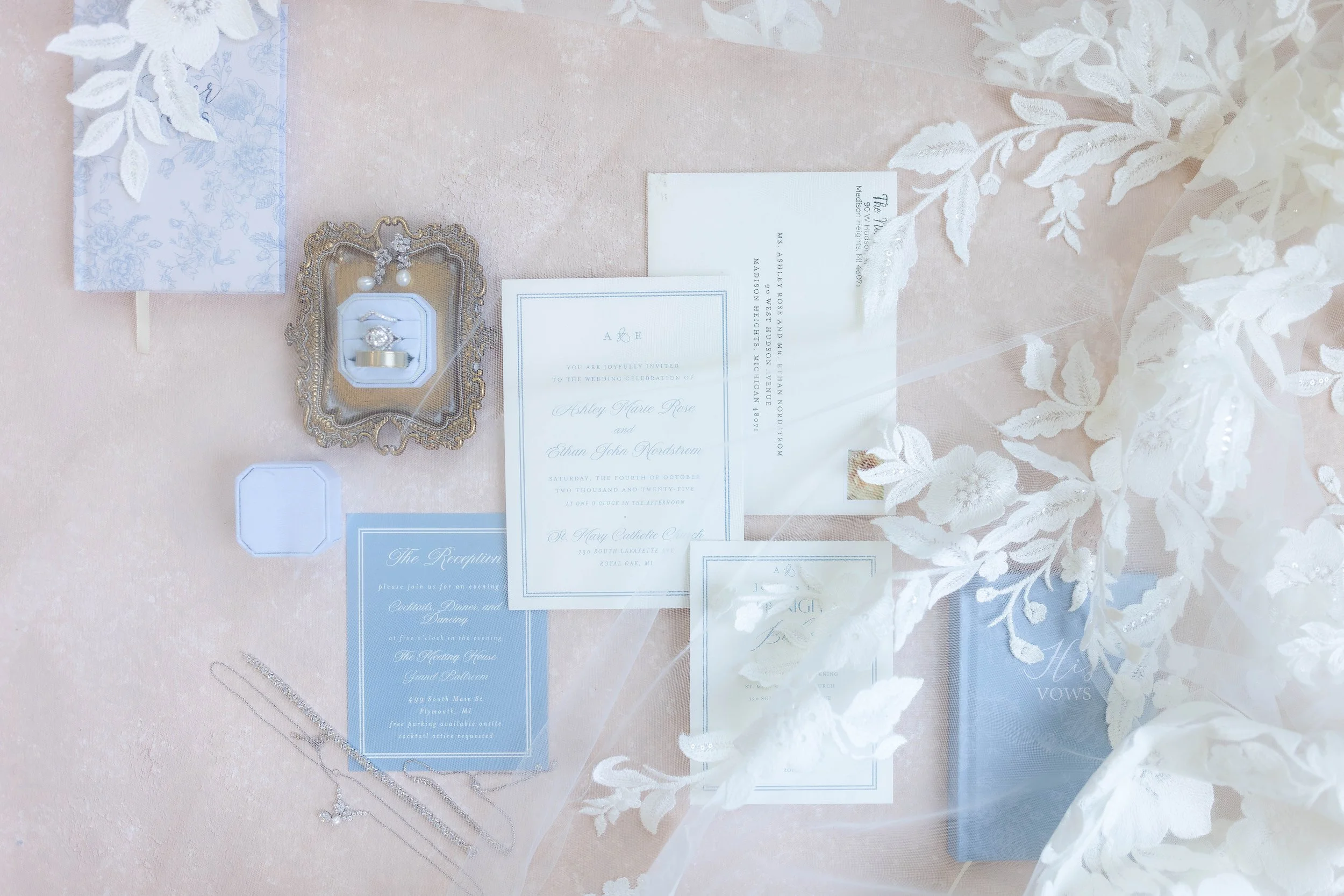 Wedding invitation suite including a main invitation card, reception card, and other stationery, decorated with white lace and floral accents, with jewelry and delicate accessories nearby, set on a pink fabric background.