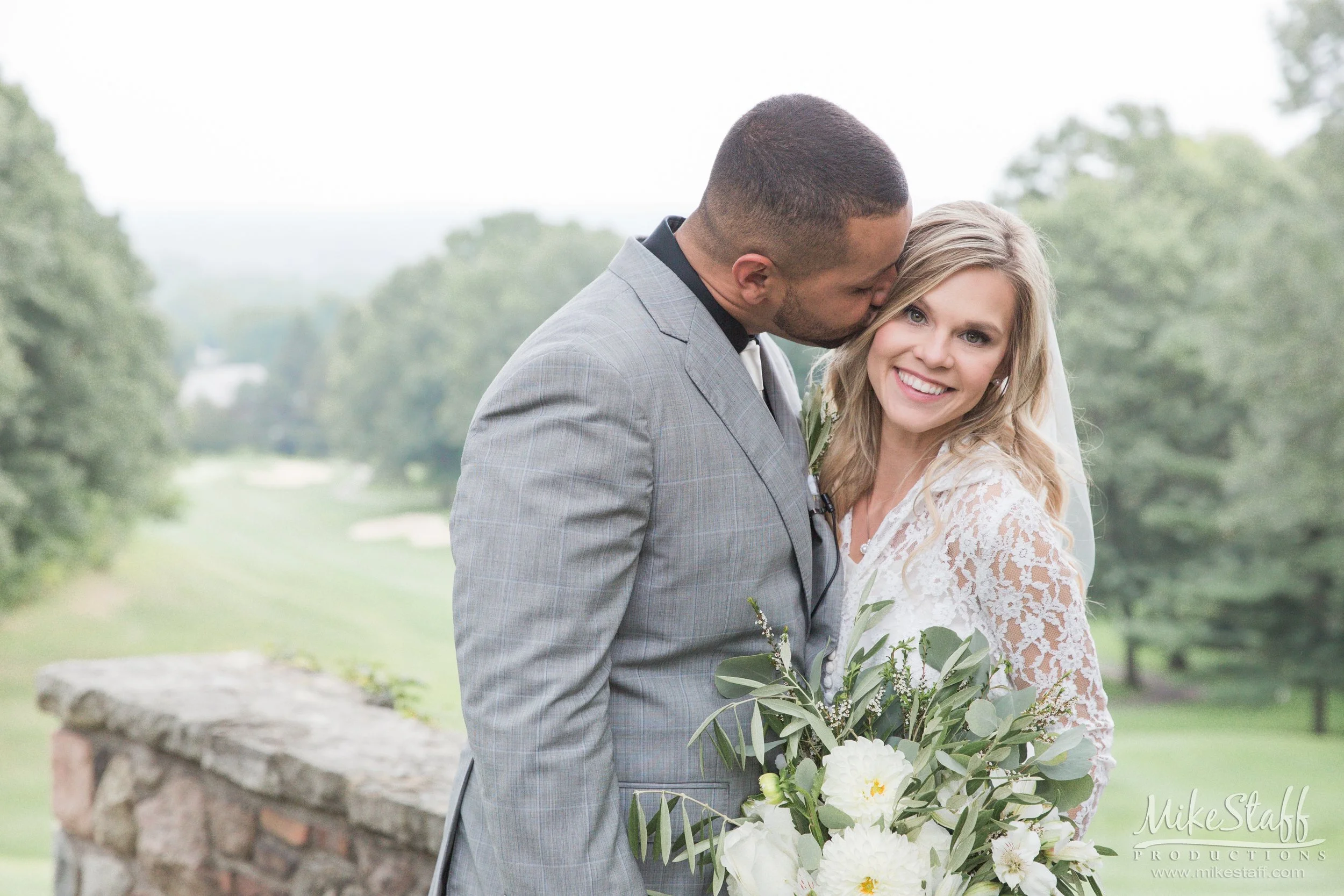 A man dressed in a gray plaid suit kisses a smiling woman in a white lace dress on her temple during a wedding ceremony outdoors. The woman holds a bouquet of white flowers and greenery. There are trees and a stone bench in the background.