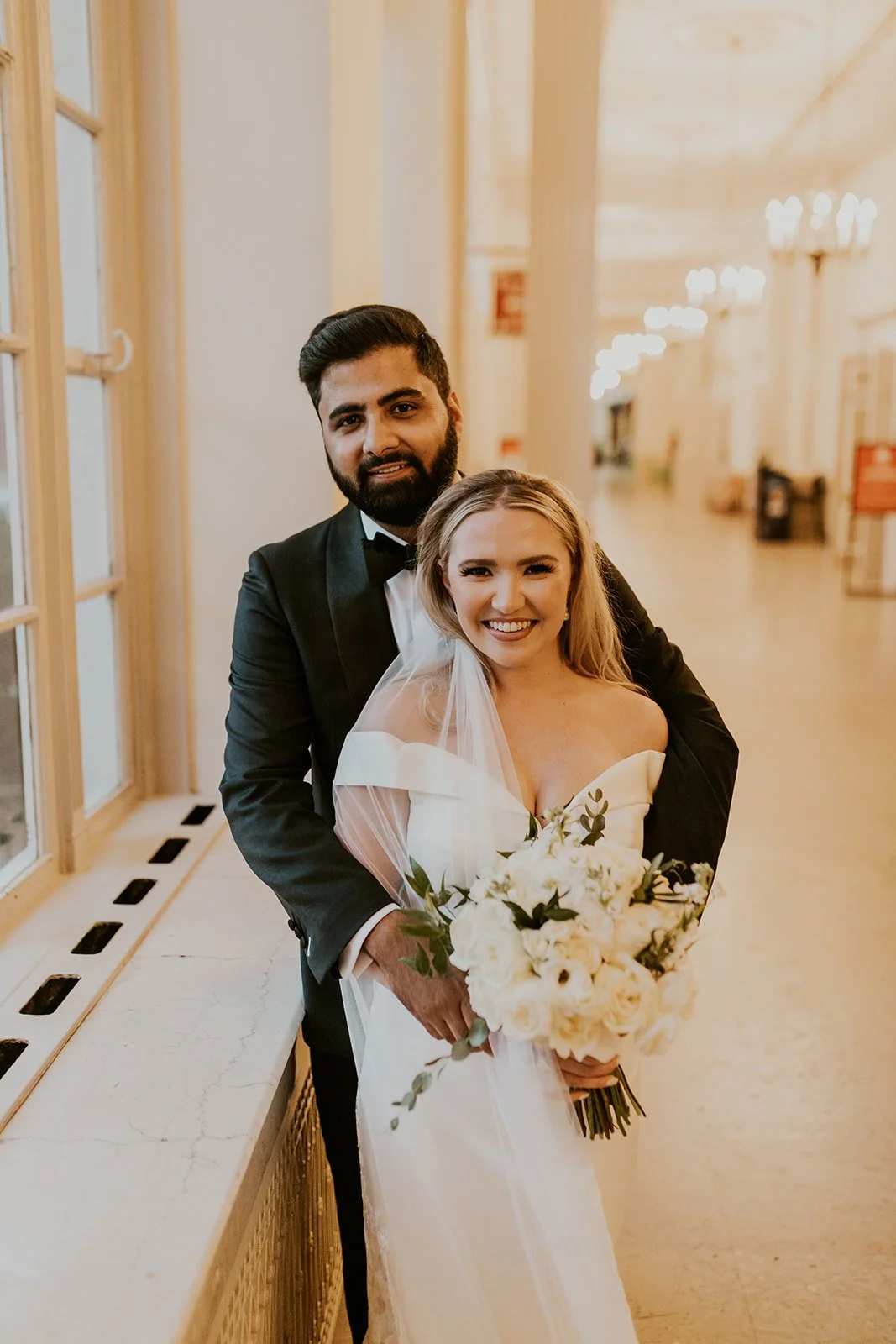 A newlywed couple standing in a hallway, the groom in a black tuxedo and the bride in a white wedding gown holding a bouquet of white flowers. They are smiling.