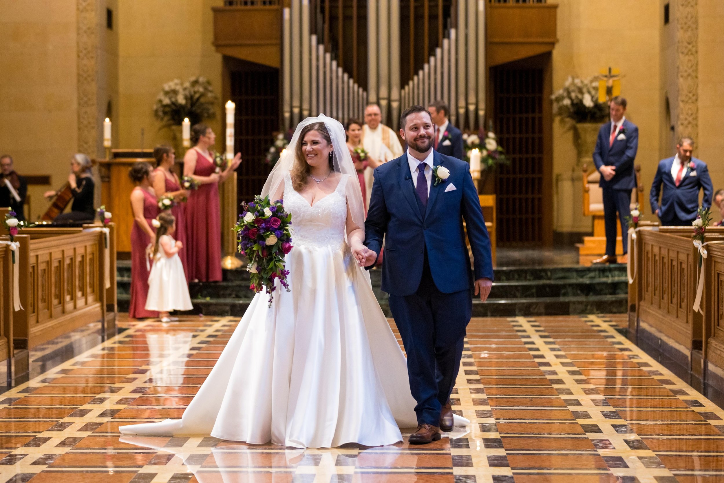 A bride and groom holding hands and smiling during their wedding ceremony inside a church, with wedding party members standing on either side and choir members softly clapping in the background.